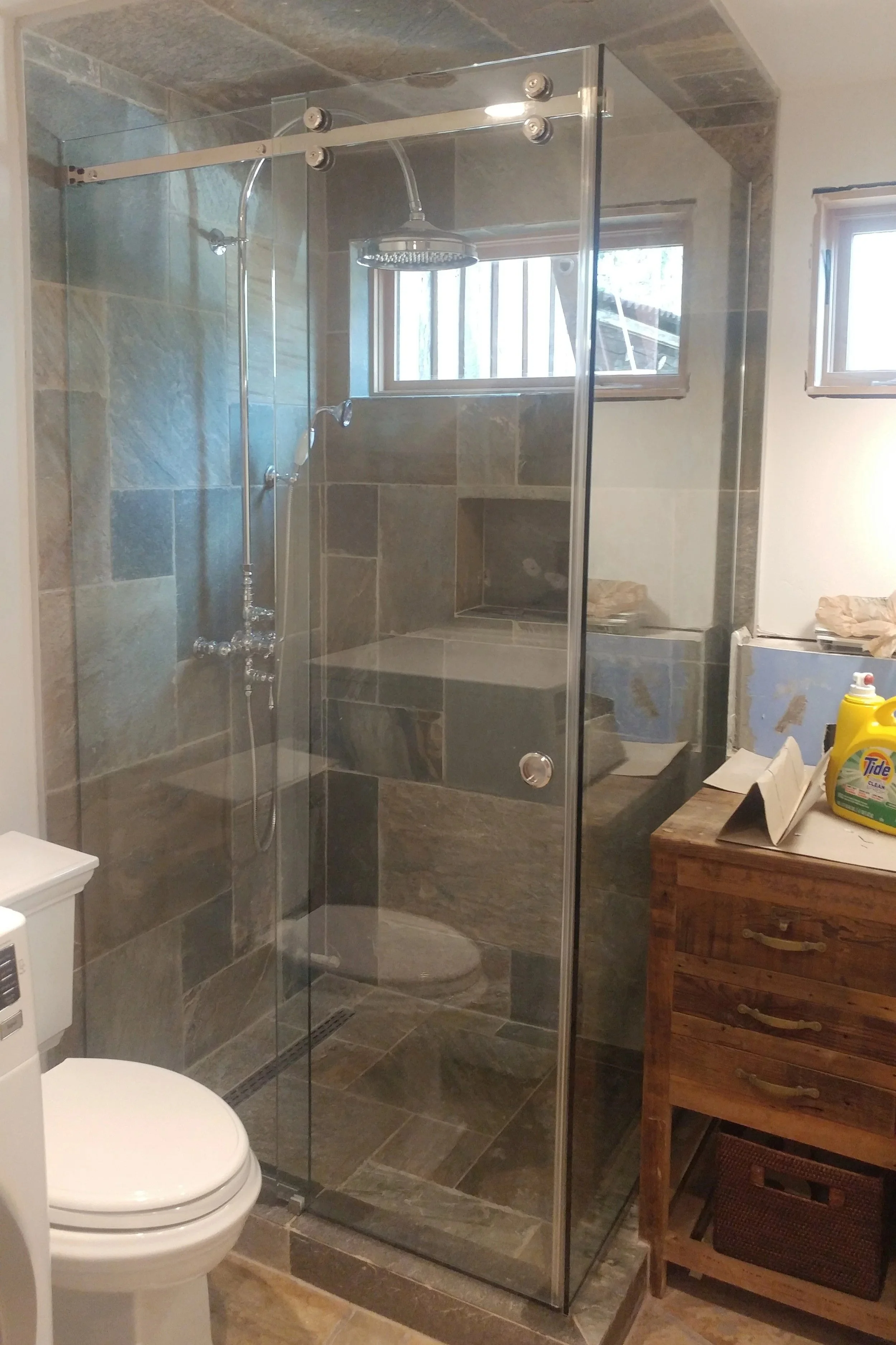 A bathroom with a glass-enclosed shower featuring gray and tan stone tiles, a rainfall showerhead, and a window with wooden blinds. There is a toilet in the foreground and a wooden cabinet with various items on top to the right.