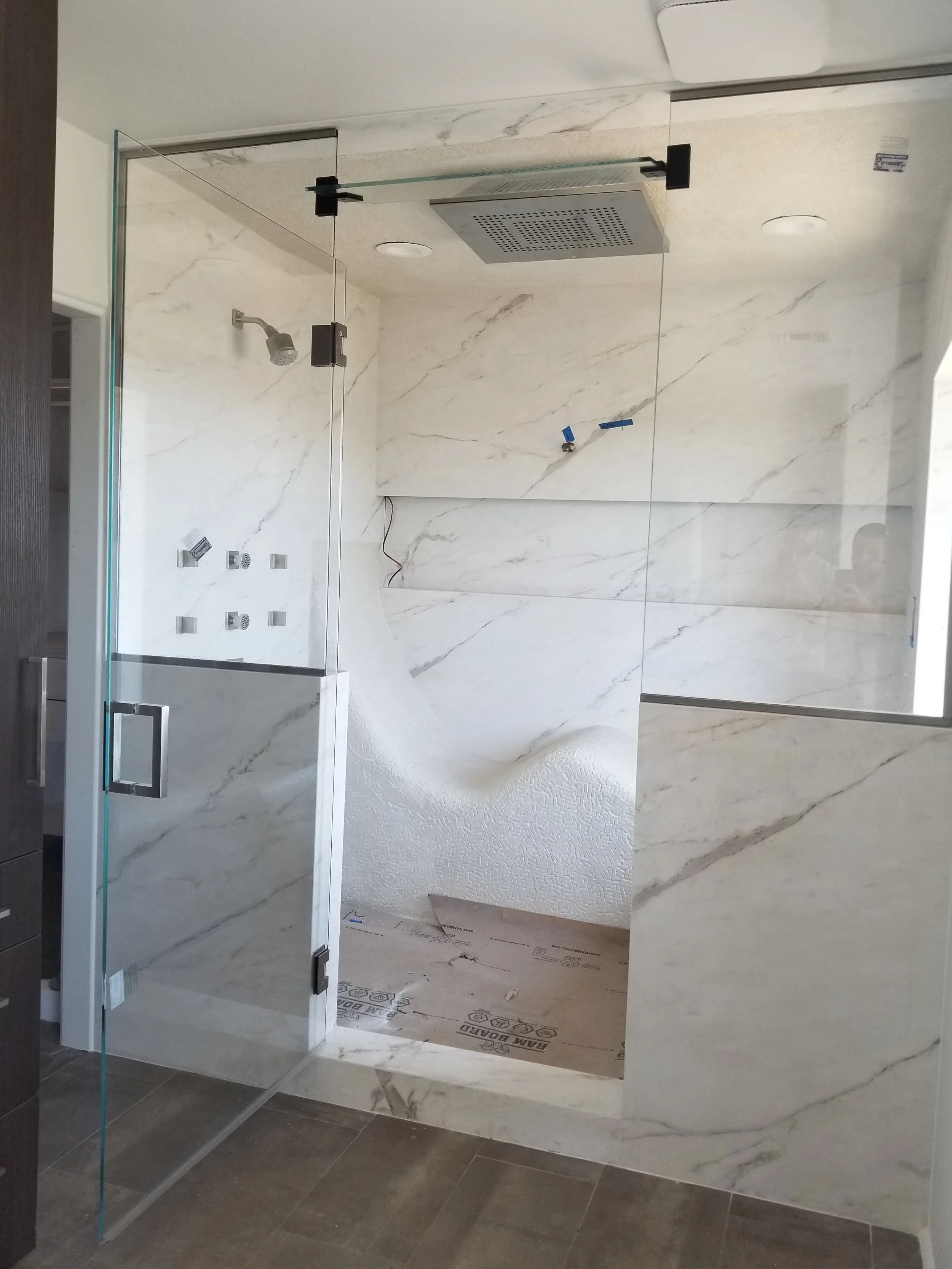 Empty bathroom with marble walls and floor, glass shower enclosure with black hinges, large showerhead, and recessed lighting.