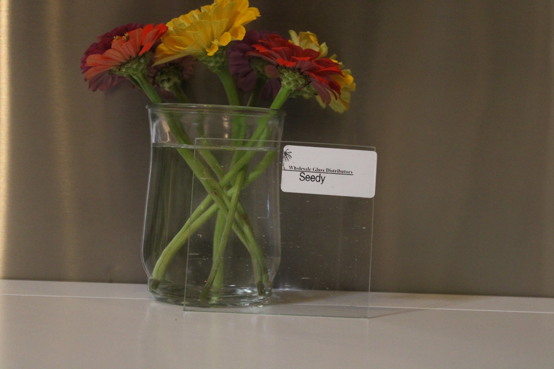 A clear glass vase containing a bouquet of colorful flowers, including red, yellow, and purple blossoms, placed on a surface against a plain background with a label reading "Whole sale Glass Distributors Seedy".