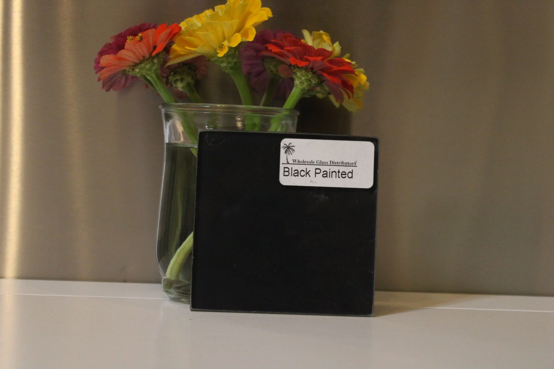 A clear glass vase with pink, yellow, and red flowers next to a black sign that says "Wholesale Glass Distributors" and "Black Painted."
