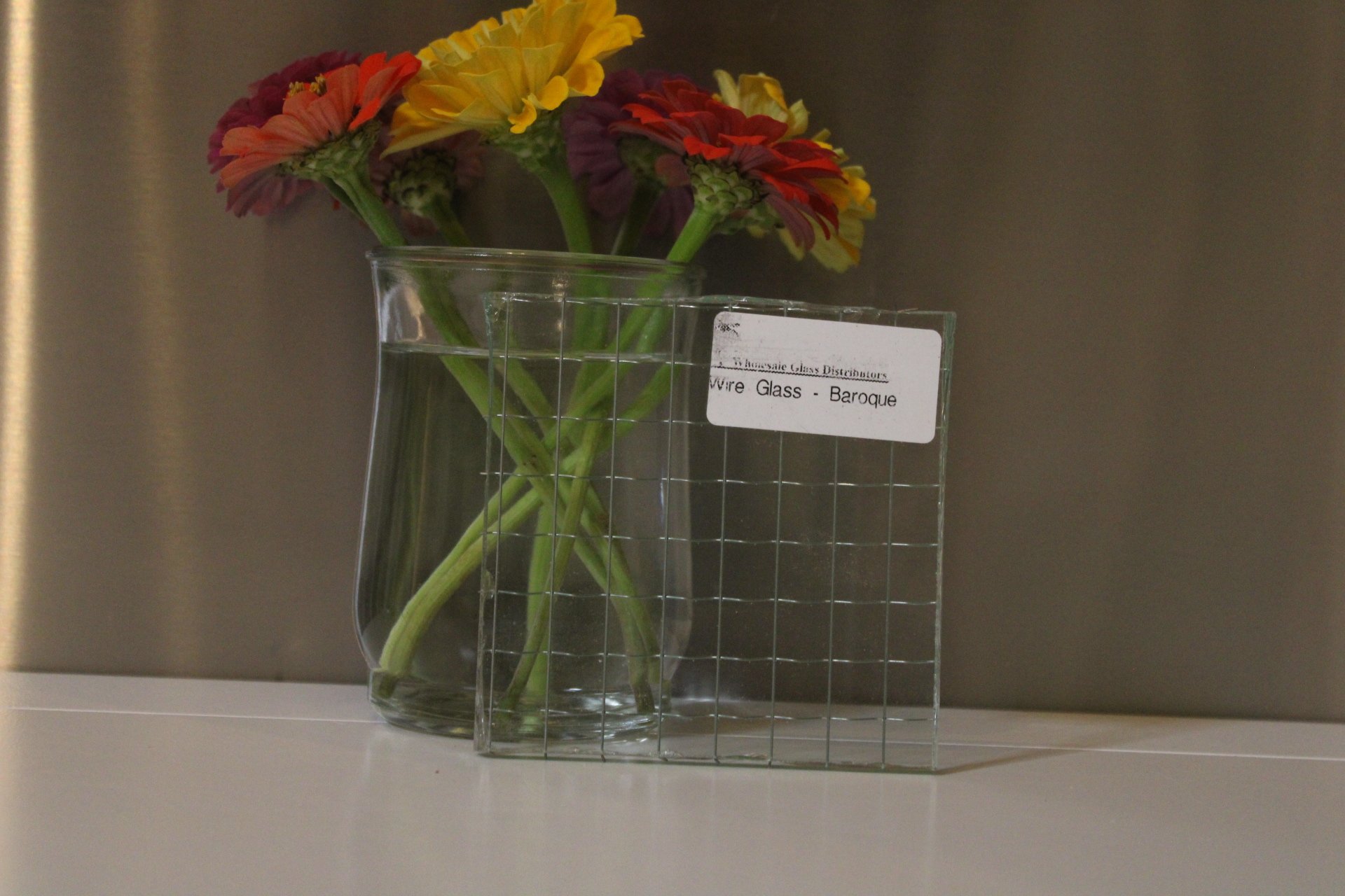 A clear glass vase with a wide opening, holding colorful flowers including yellow, orange, and red blooms with green stems, placed on a white surface against a gray background. There is a sticky note on the vase that says 'Wire Glass - Baroque'.
