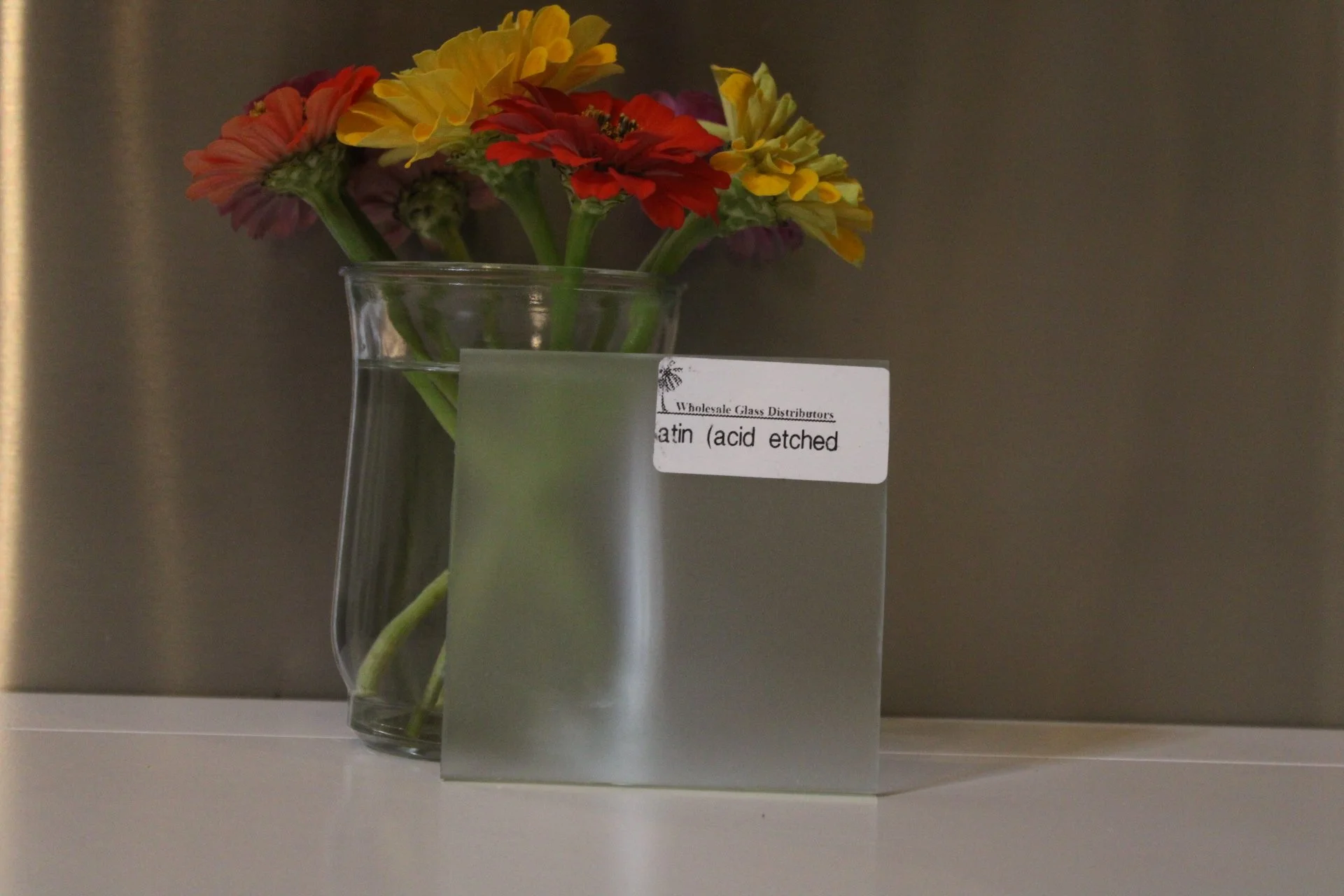 A clear glass vase with colorful flowers, including red, yellow, purple, and orange blooms, on a white surface. There is a label on the vase reading 'Wholesale Glass Distributors Satin (acid etched)...' and some blurred text.
