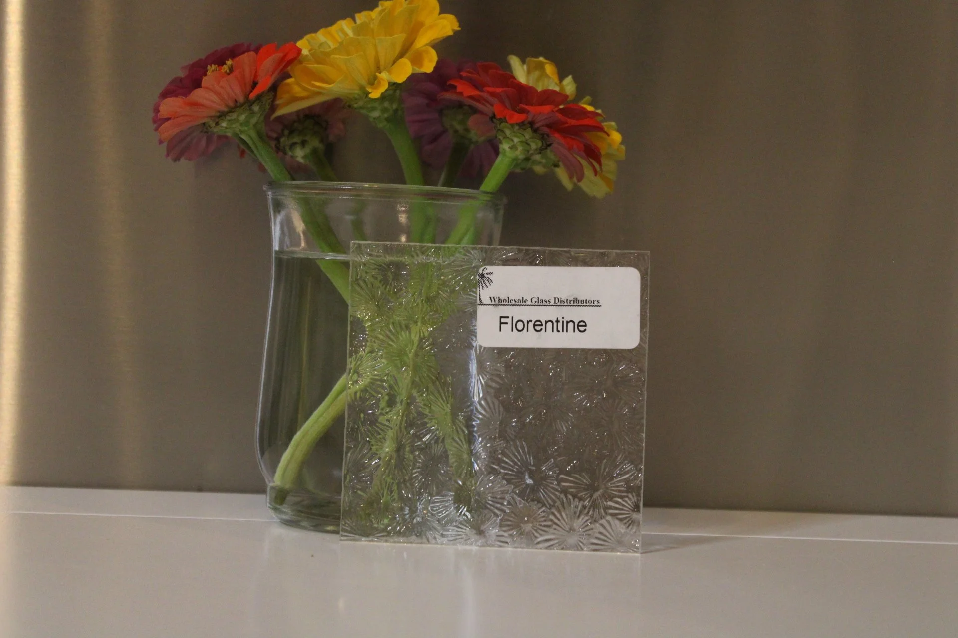 A clear glass vase with colorful flowers including yellow, orange, red, and purple blossoms, some placed inside a transparent decorative glass cube displaying the word "Florentine" and the text "Wholesale Glass Distributors".