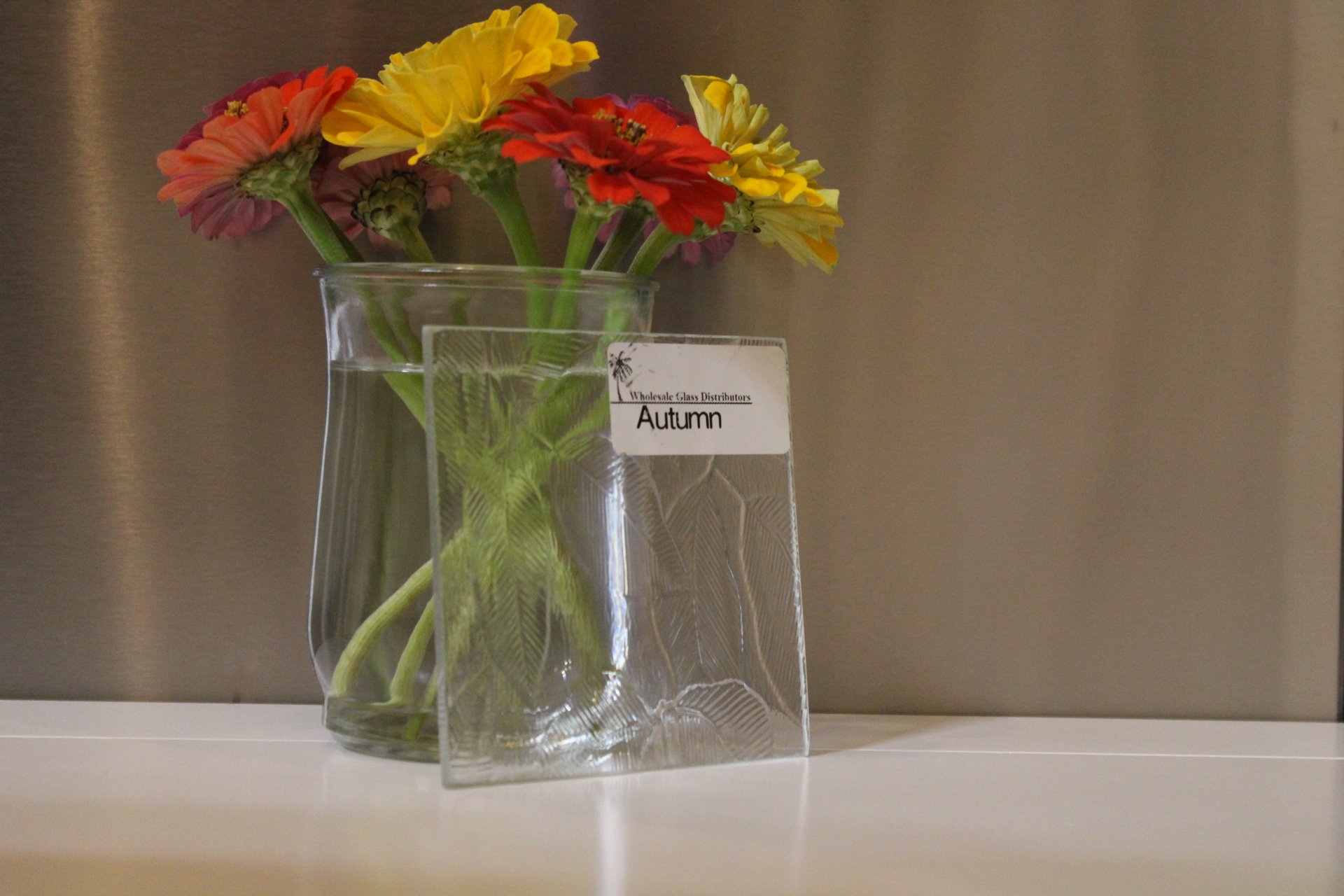A glass vase with water holds colorful flowers including yellow, red, and pink, labeled 'Autumn' from a wholesale glass distributor. The vase is placed on a white surface against a neutral-colored background.