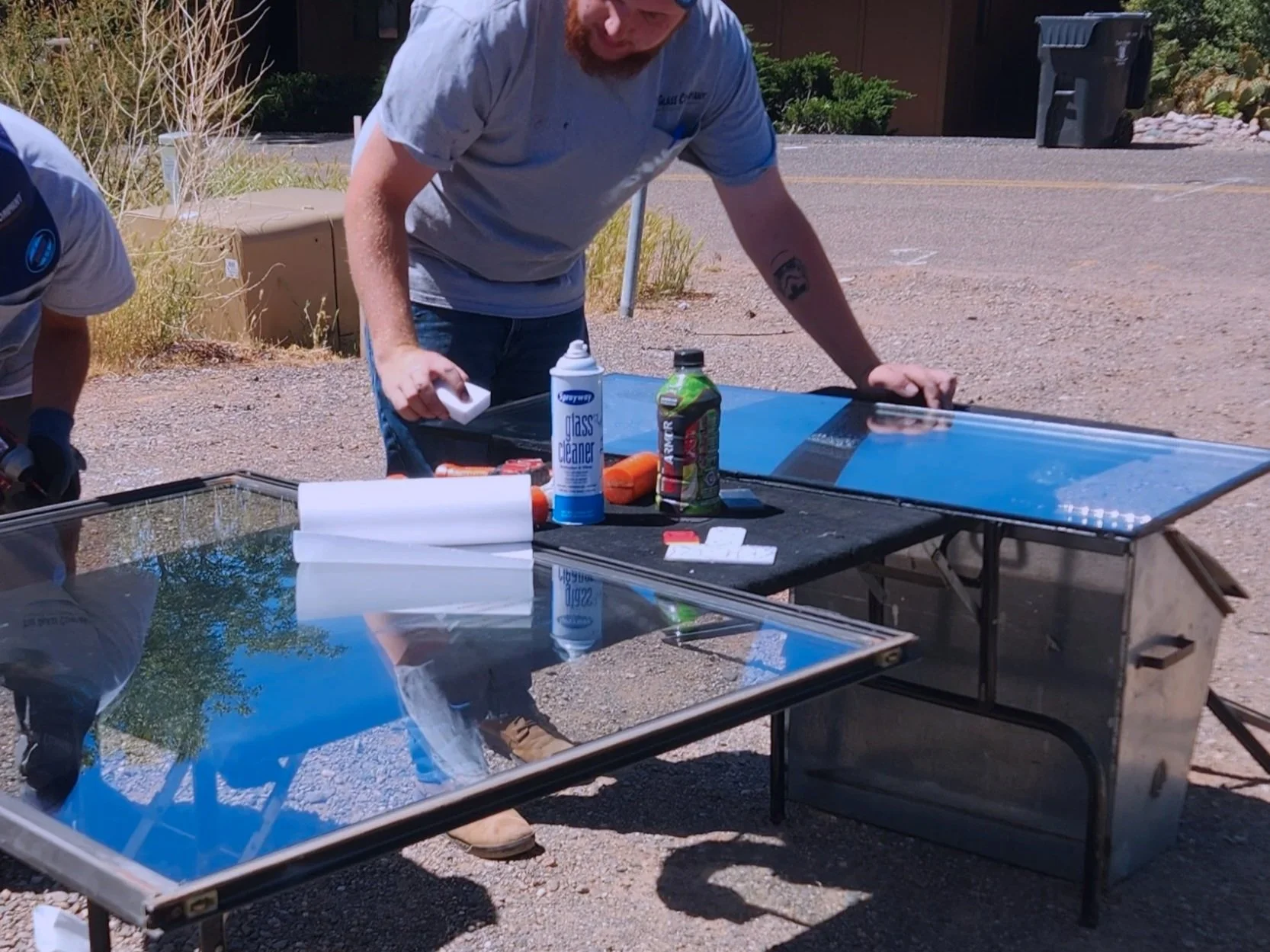 Man cleaning glass screen on outdoor table with cleaning spray, paper towels, and a spray bottle; another person partially visible on left; background includes bushes and a trash bin.