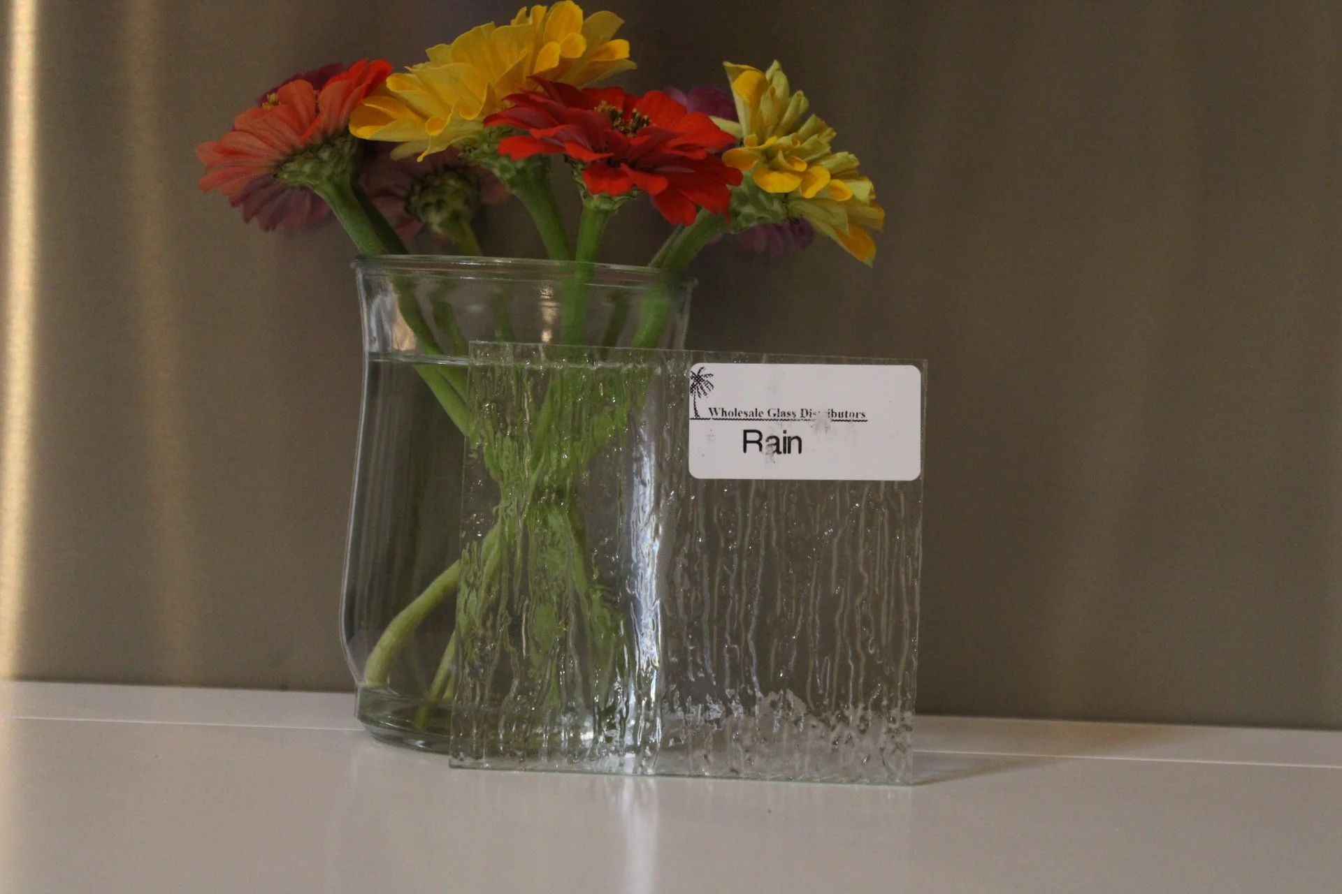 Colorful flowers in a clear glass vase on display, labeled 'Rain' from Wholesale Glass Distributors.