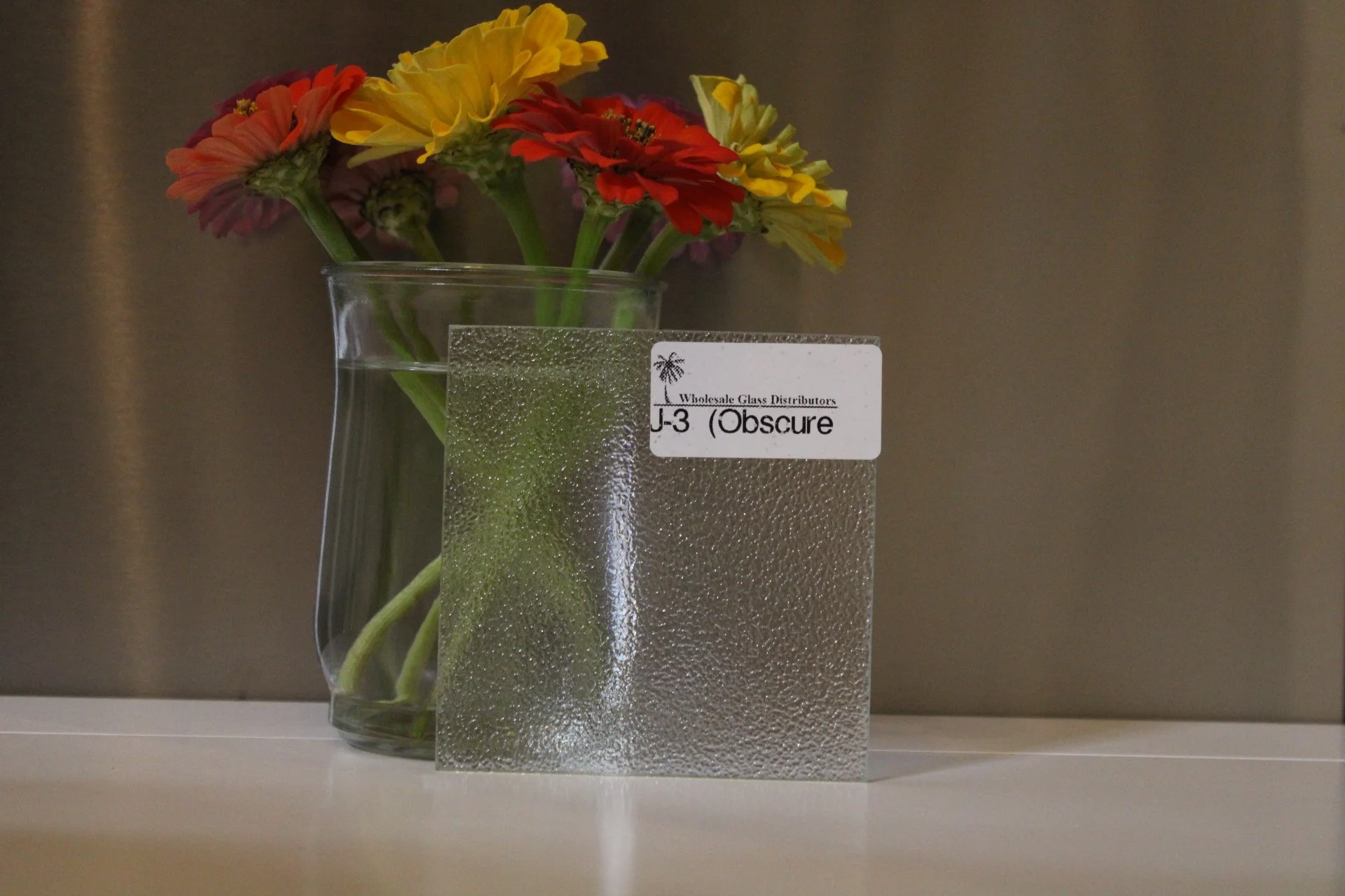 A clear glass vase with water, holding a bouquet of yellow and red flowers, partially obscured by a textured glass sheet with a label that reads 'Wholesale Glass Distributors J-3 (Obscure)'. The background is a plain gray wall.