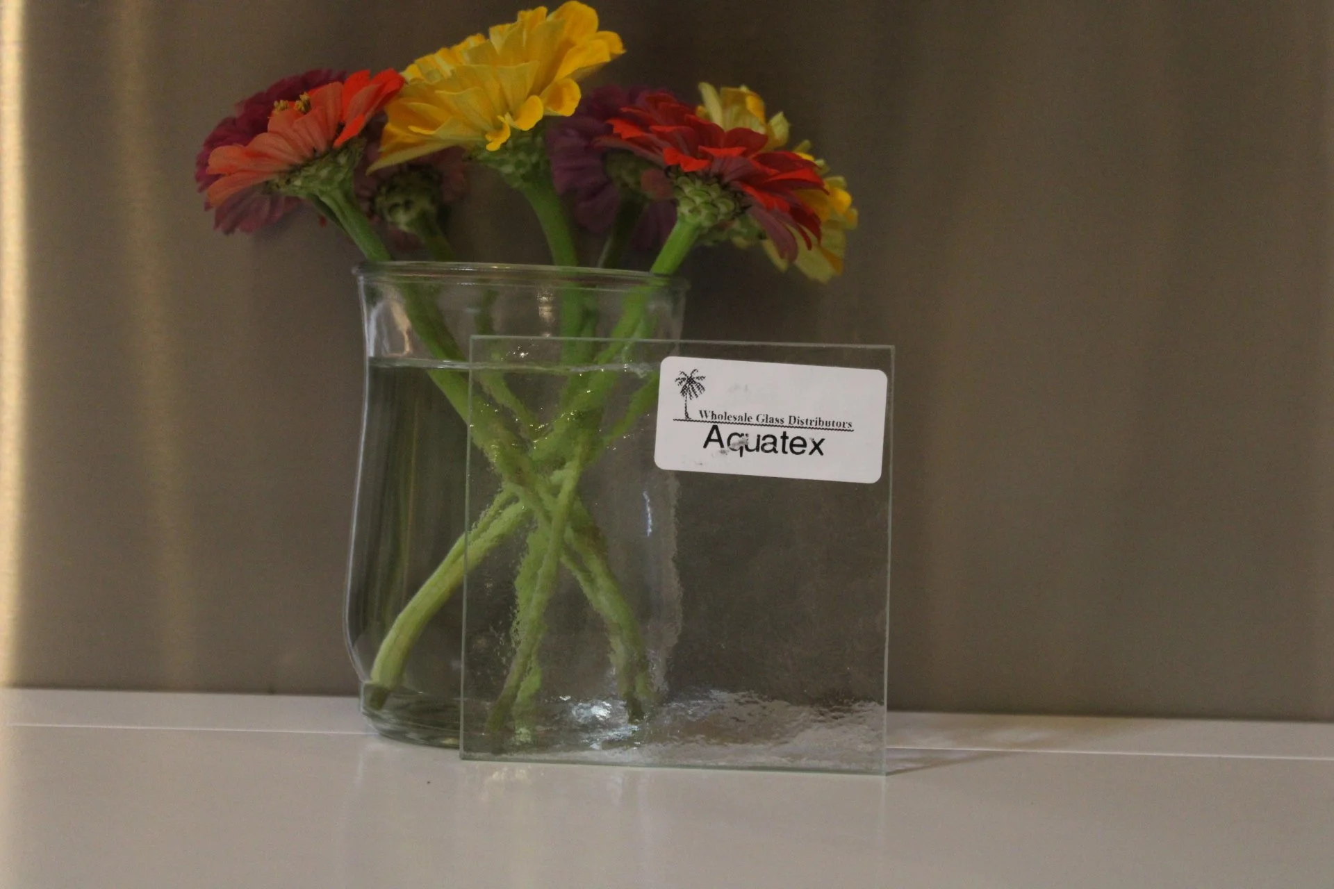 A glass vase with colorful flowers, including yellow and red zinnias, on a white surface against a neutral background, with a label reading 'Wholesale Glass Distributors Aquatex'.