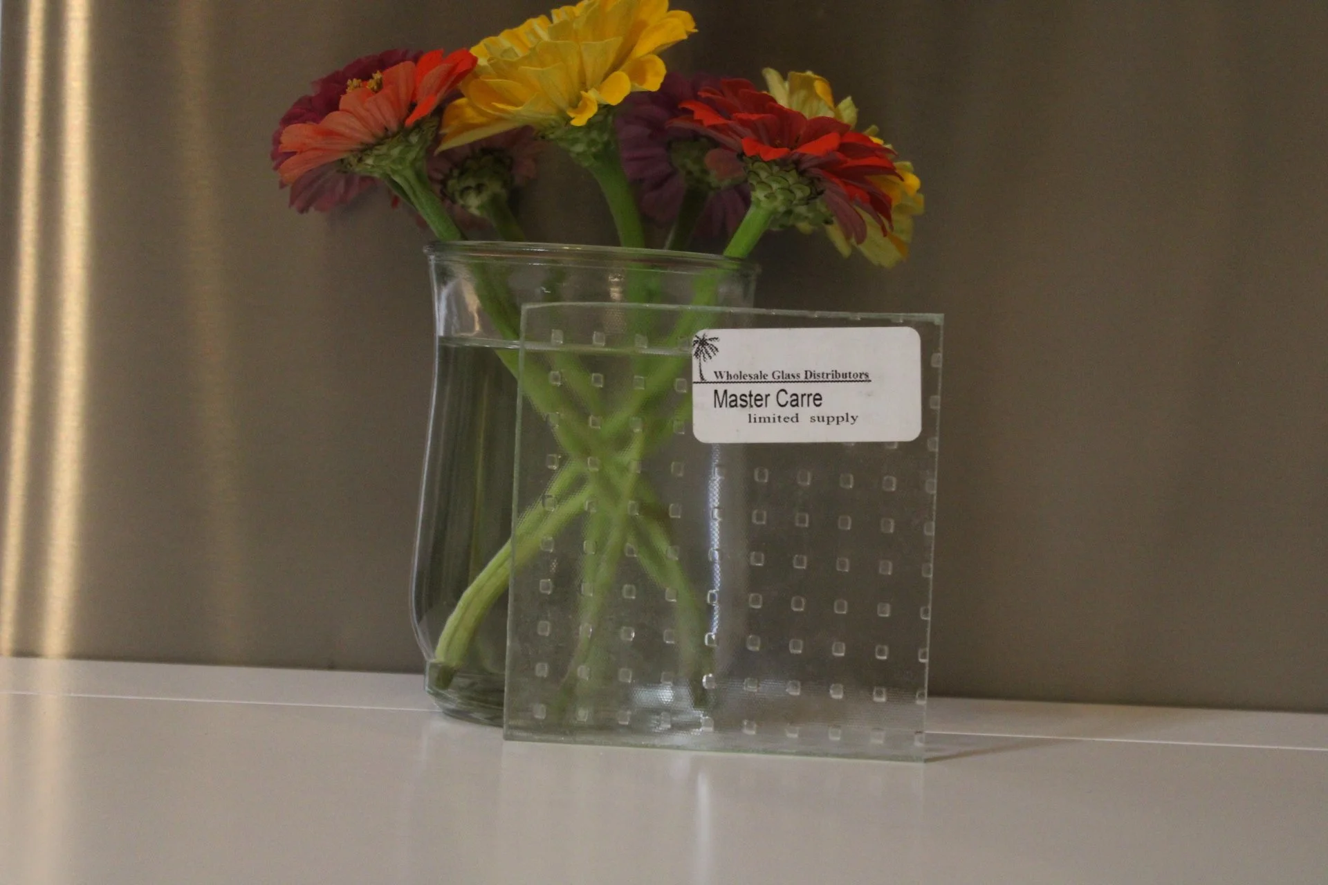 Colorful flowers in a glass vase on a countertop with a sign that reads 'Wholesale Glass Distributors, Master Care, limited supply'.