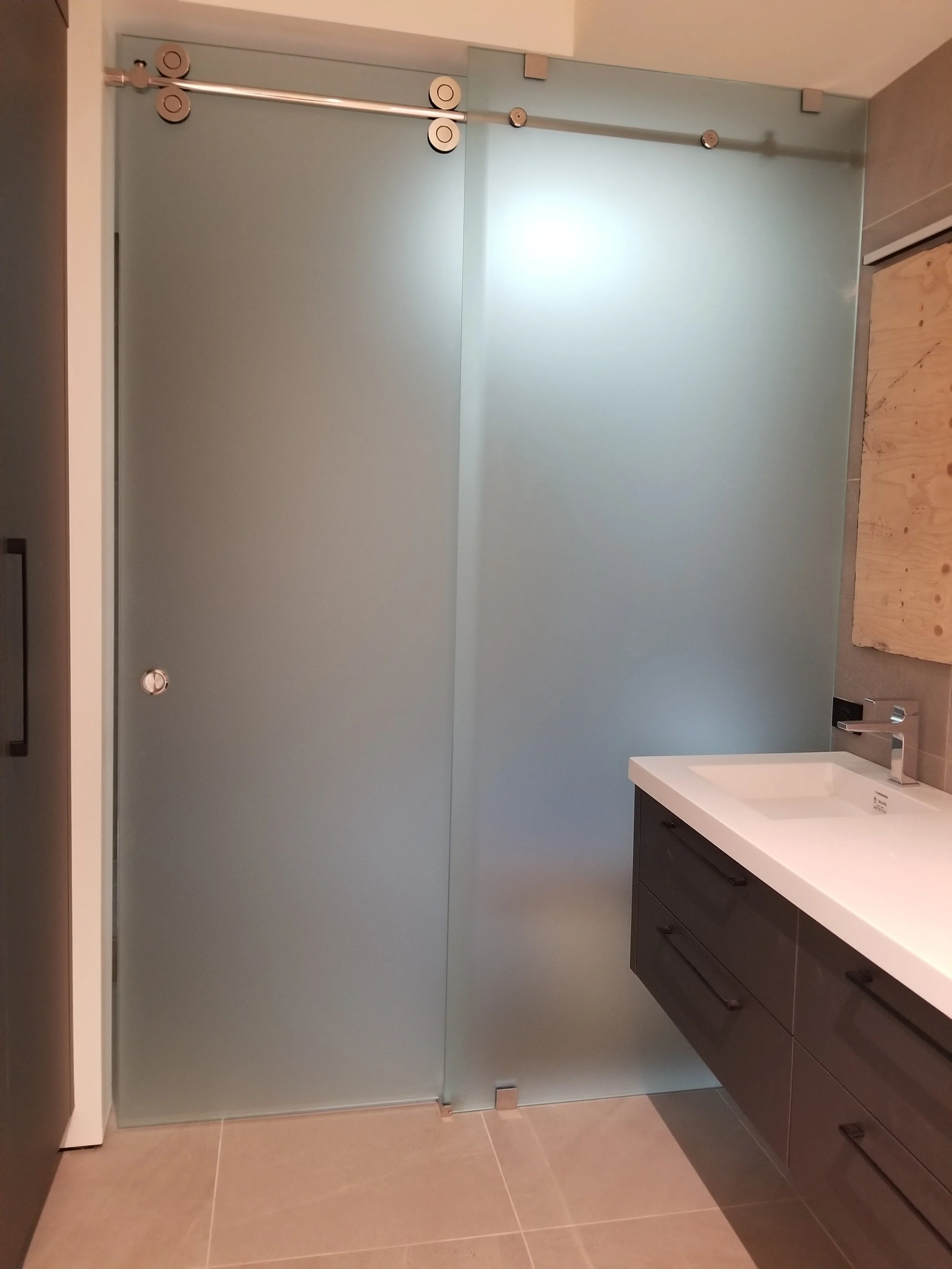 A modern bathroom with a frosted glass shower door on the left and a dark wood vanity with a white sink on the right. The floor is tiled with beige tiles.
