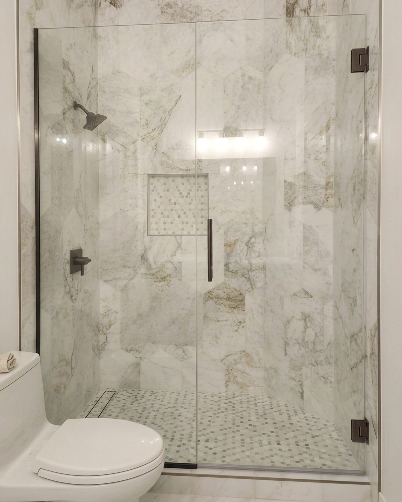 A bathroom with a glass-enclosed shower featuring marble walls and a built-in shower niche. A white toilet is partially visible in the lower left corner.