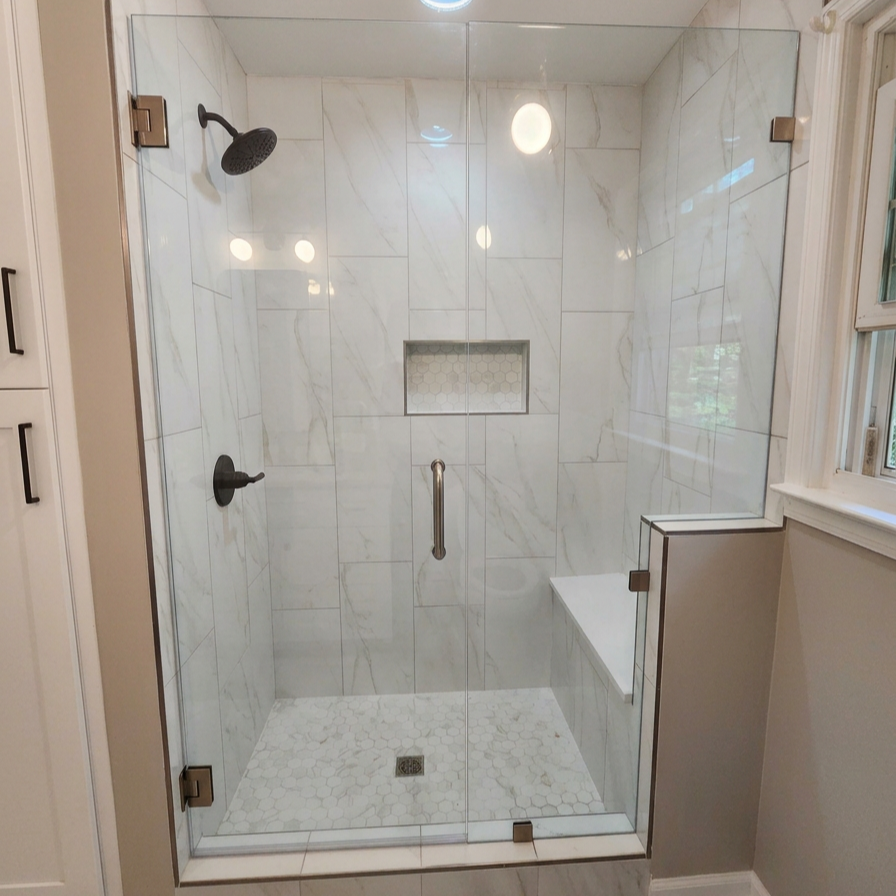 A modern walk-in shower with white marble tiles, a black showerhead, and black control handles. There is a built-in shelf with a hexagonal tile pattern, and a bench with a white surface and black border. The shower has a glass door with metal hinges and a handle. A window with white framing is next to the shower.