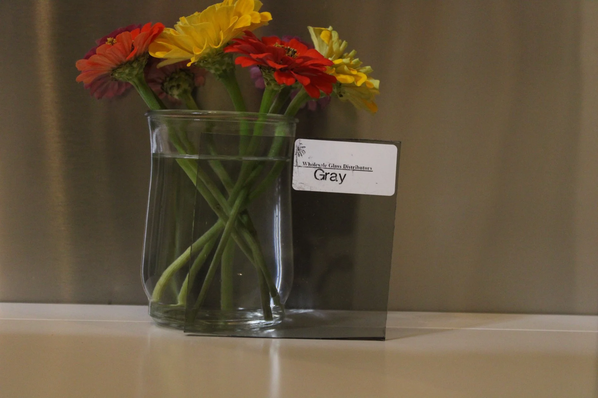 A clear glass vase with water holding a bouquet of colorful flowers including yellow, red, and pink blossoms, placed on a shelf with a gray wall background. There is a white label on the vase that says "Wholesale Glass Distributors Gray."