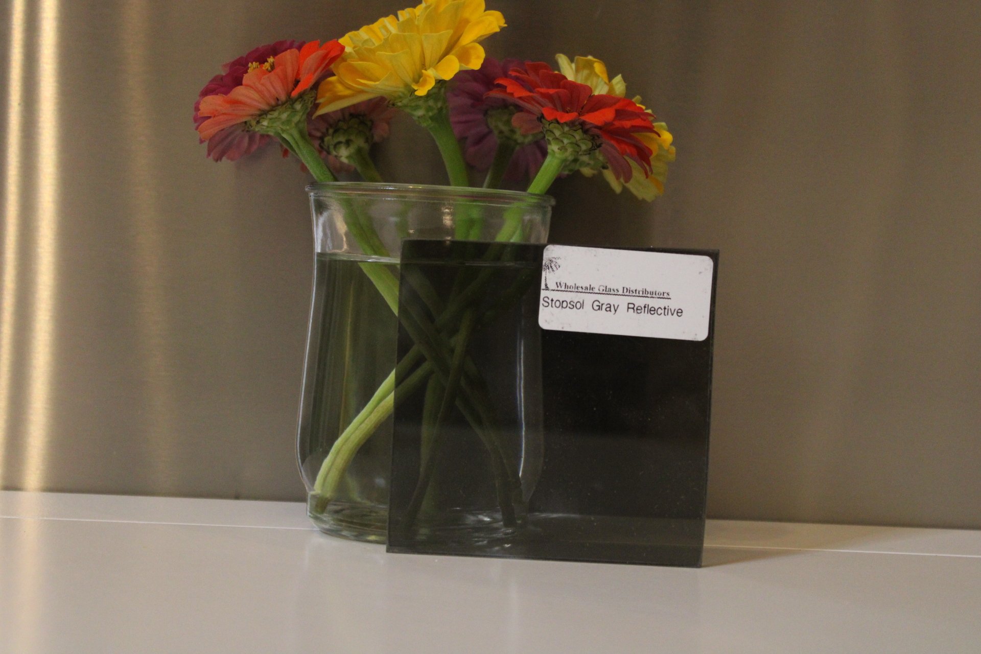 A clear glass vase with a bouquet of colorful flowers containing yellow, orange, and purple blooms. A black card with white text is positioned behind the vase.