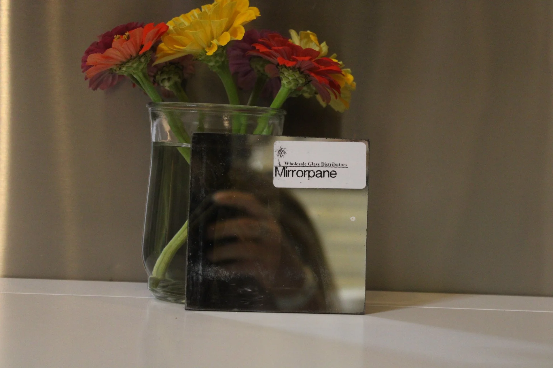 A clear glass vase with a label that says "Mirrorpane," holding a bouquet of colorful flowers including yellow, orange, and purple blossoms, placed on a white surface against a plain dark background.