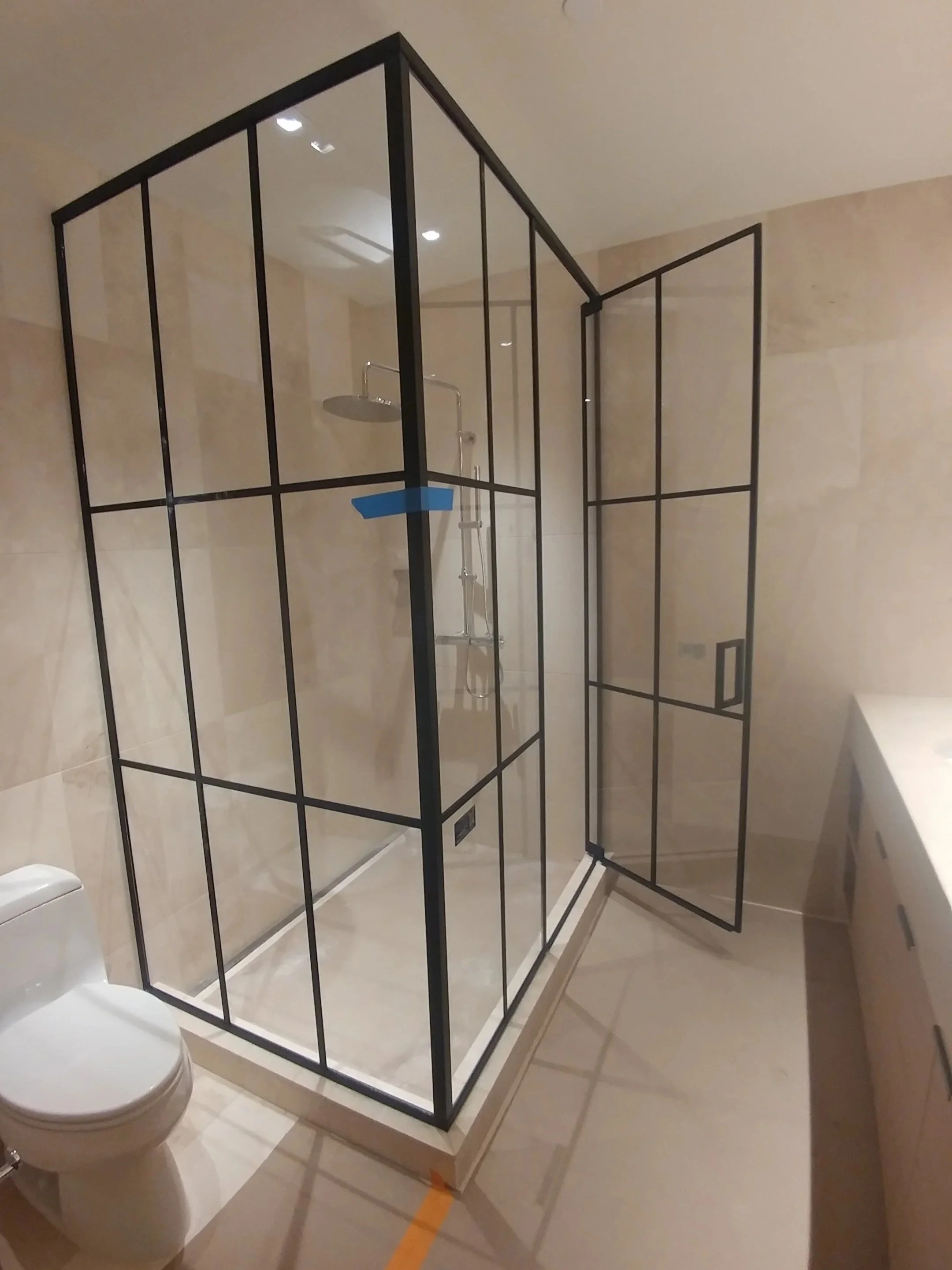 A bathroom with beige tile walls and floor. A black metal-framed glass shower enclosure with a door is in the corner. Inside the shower, there is a wall-mounted showerhead and a handheld showerhead on a metal pole. A white toilet is partially visible in front of the shower.