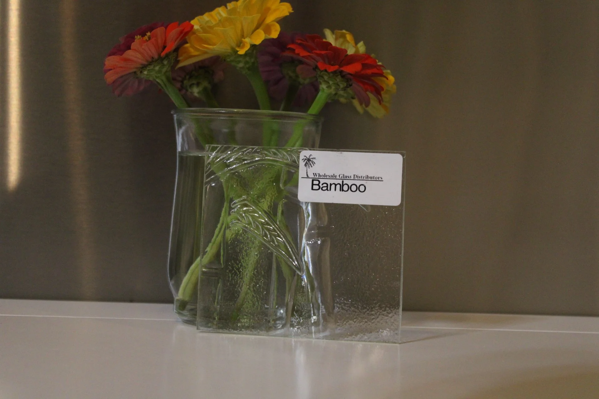 A glass vase with an intricate leaf design contains a few green stalks and a large leaf. Behind the vase, there are colorful flowers with yellow, red, and purple petals. There is a white label on the vase that reads: 'Wholesale Glass Distributors Bam