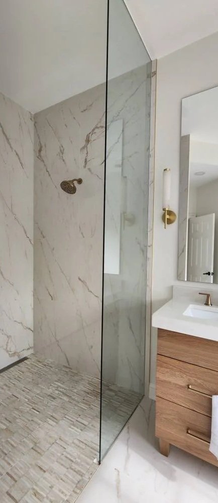 Modern bathroom shower with marble walls, glass door, wooden vanity with a white countertop, and a mirror.