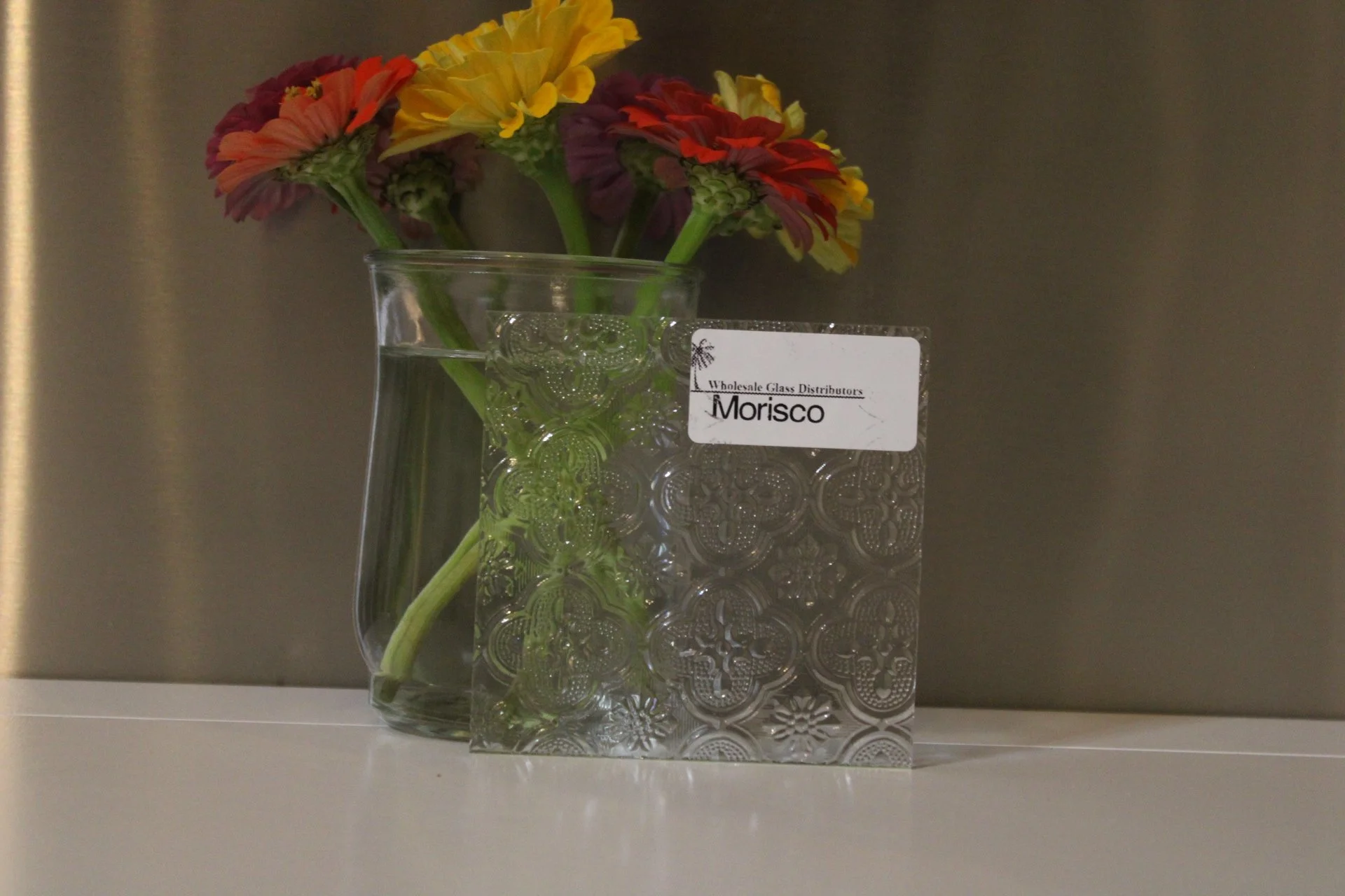 A clear glass vase with pink, yellow, and purple flowers, placed on a white surface with a decorative tissue box and a retail label that reads 'Wholesale Glass Distributors Morisco'.