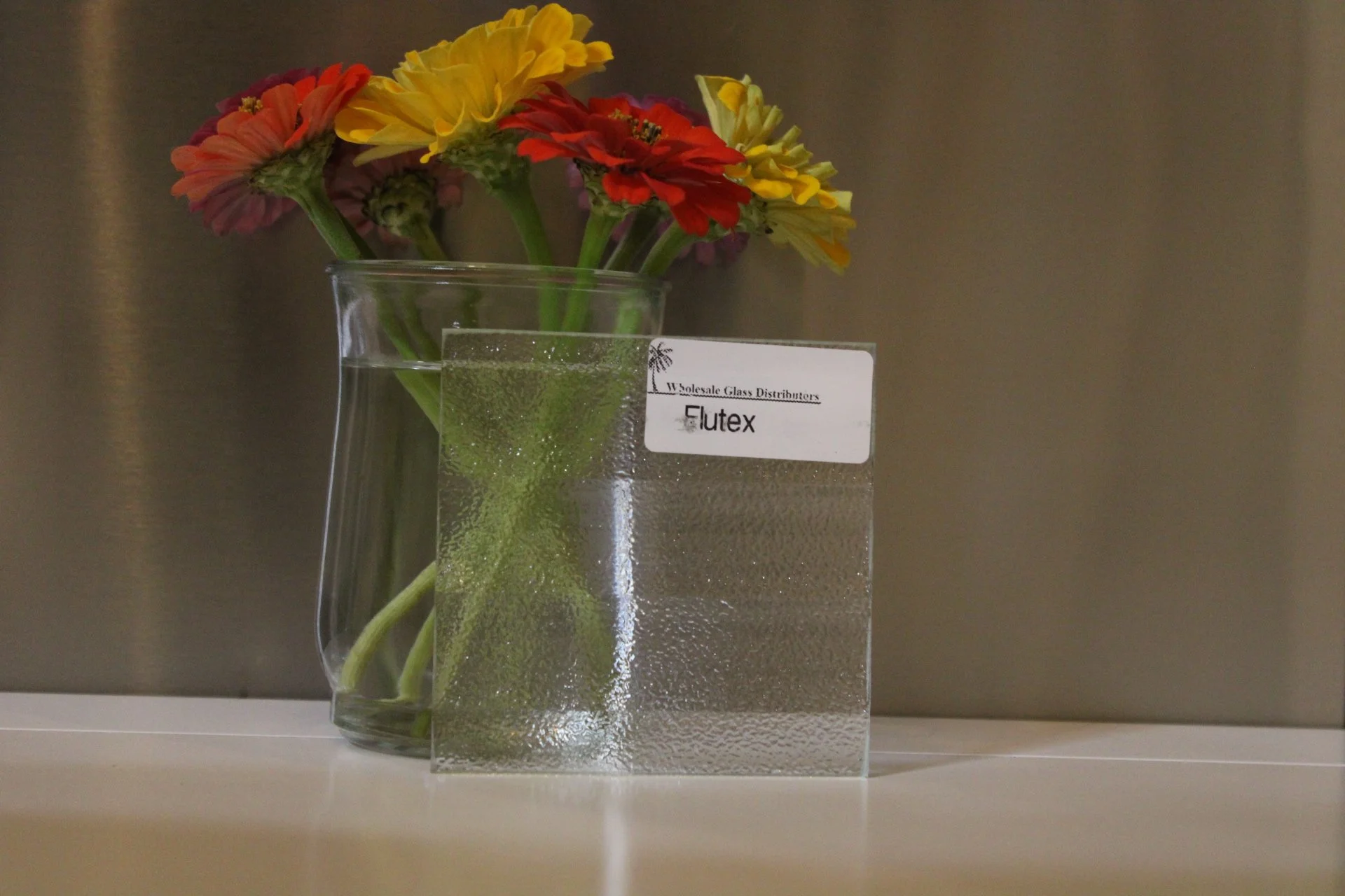 A glass vase with colorful flowers and a white card with text, placed on a white surface against a metallic background.