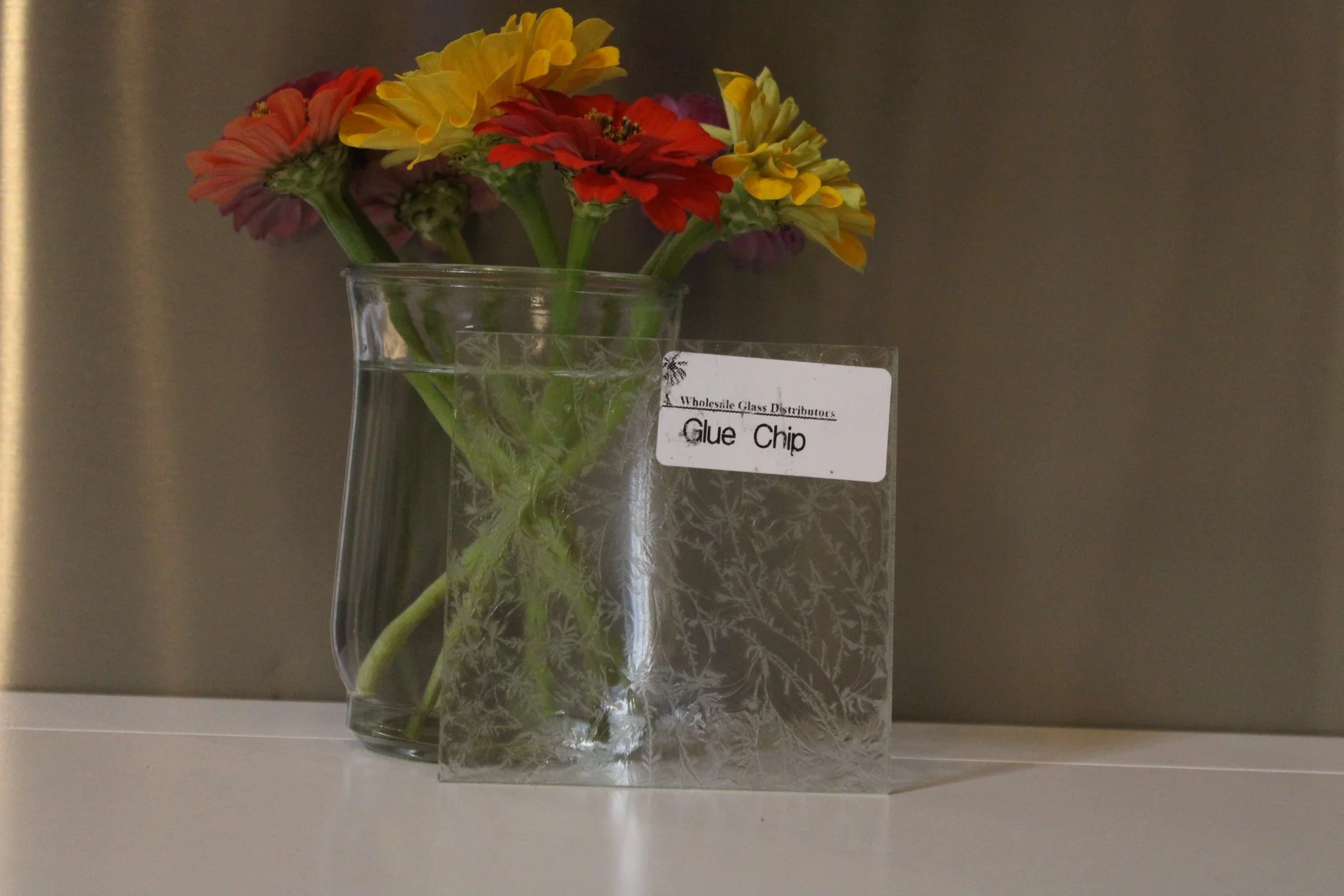 Colorful flowers in a glass vase positioned on a white surface, with a plastic label in front that reads 'Glue Chip' and a blurry background.