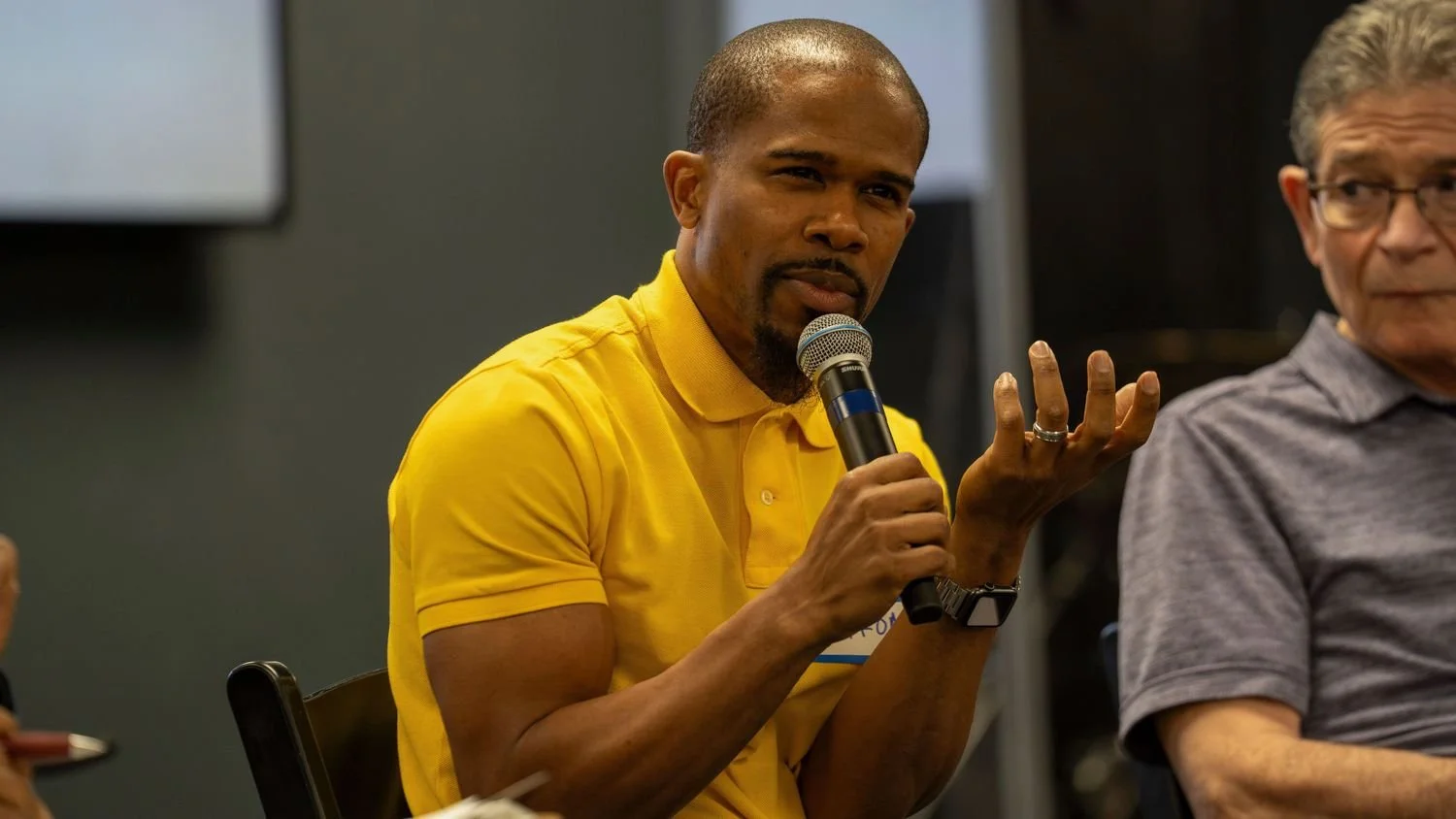 A man in a yellow polo shirt speaking into a microphone during a panel discussion, with an older man in glasses sitting beside him.