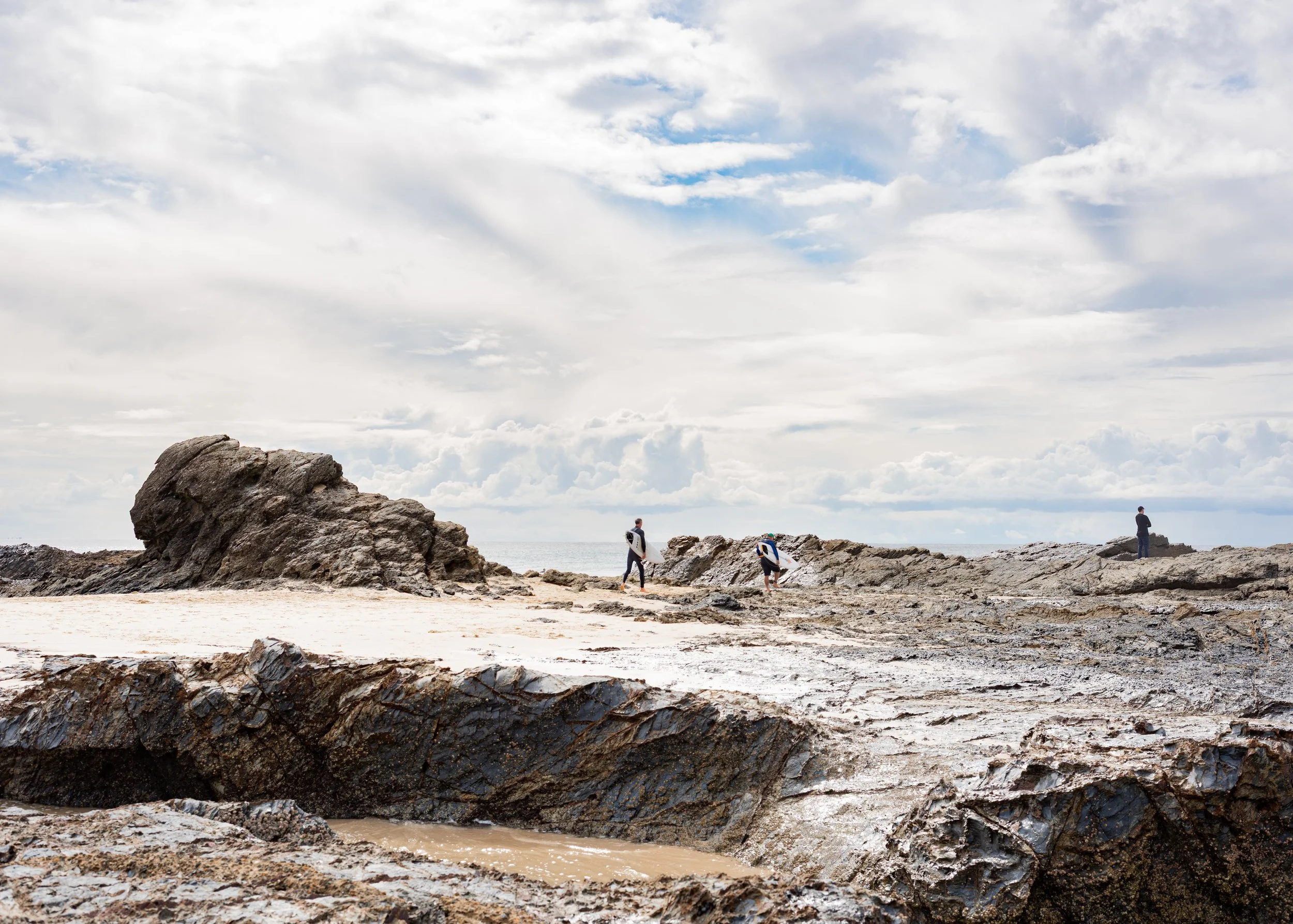 People on a rocky beach under a cloudy sky, some holding surfboards, with large rocks in the foreground and the ocean in the background.
