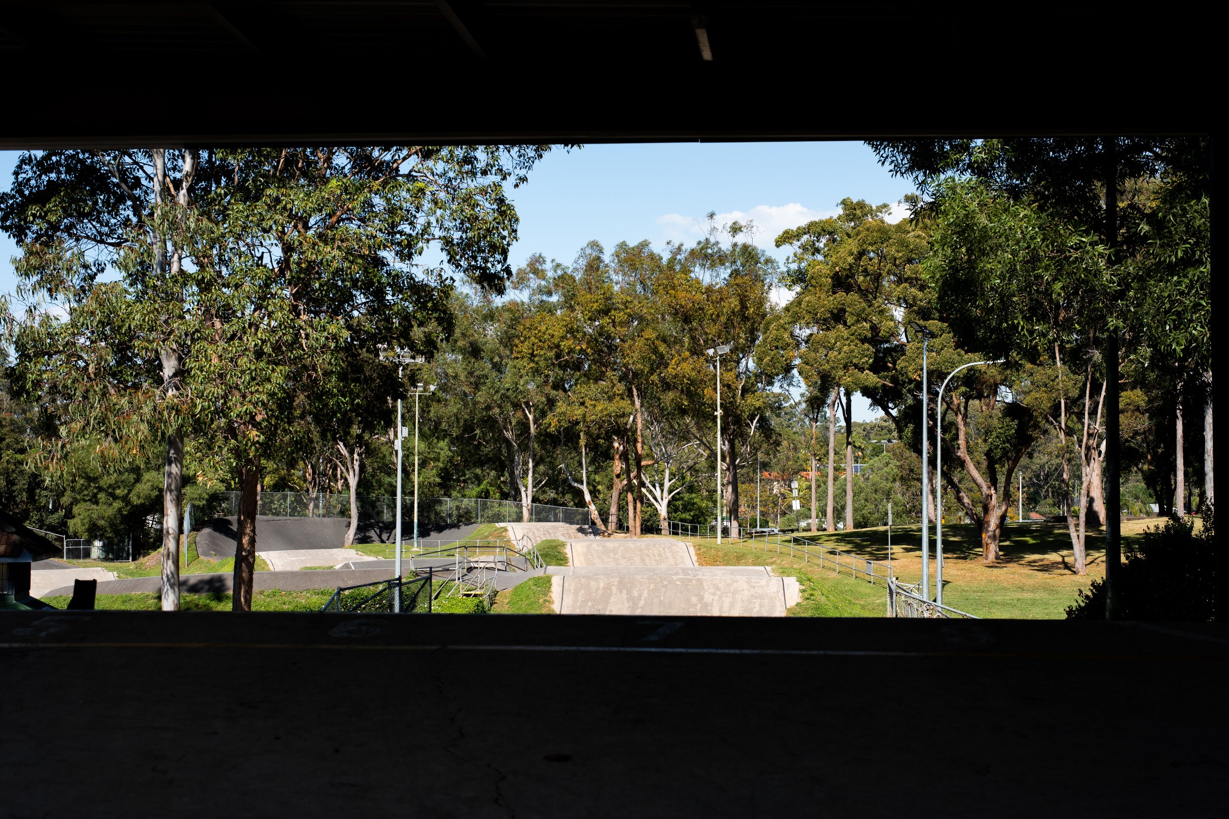 A view of a park with trees, an open grassy area, and a skate ramp, seen from a covered area with dark shadows.