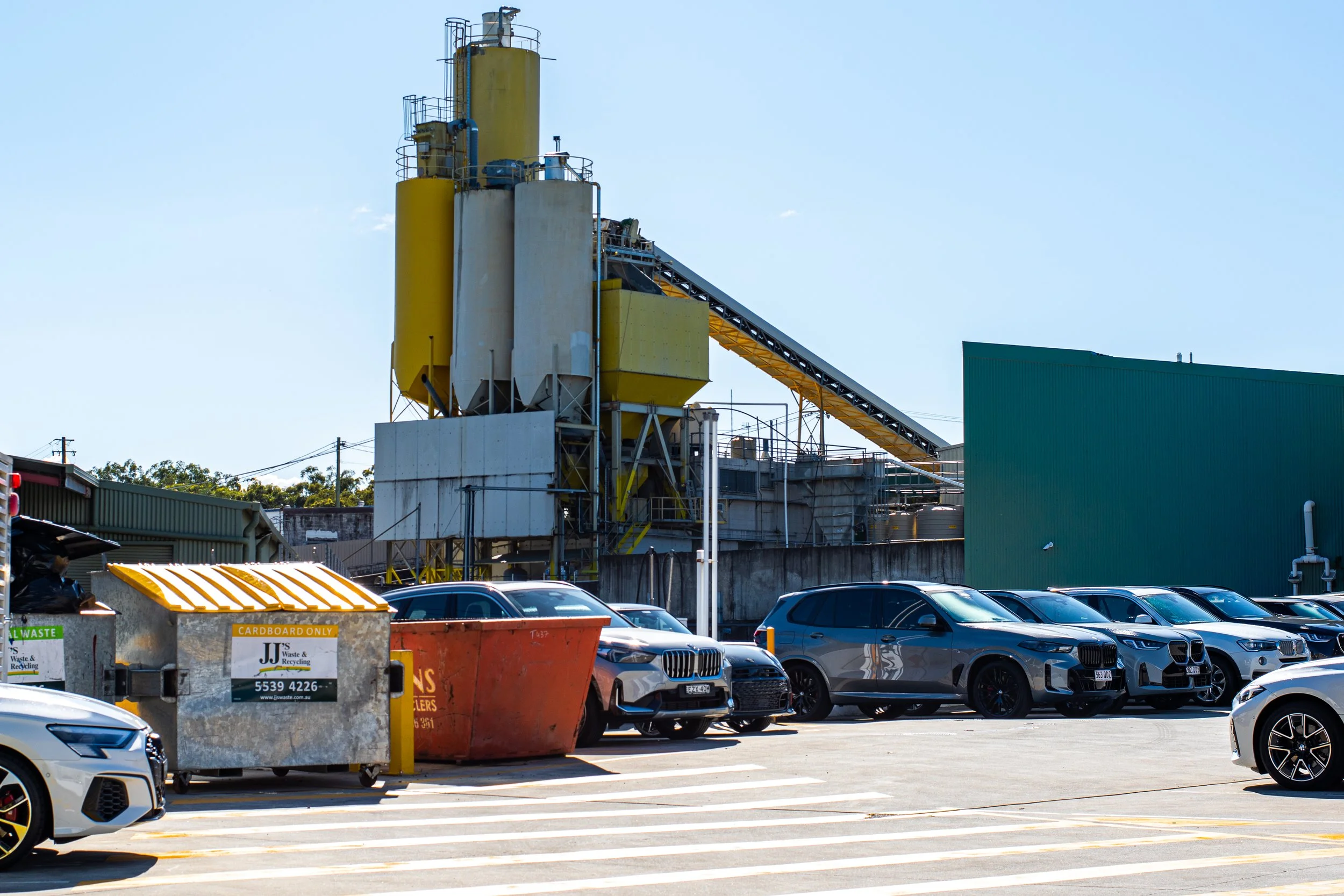 A parking lot adjacent to an industrial facility with large yellow and white silos and a conveyor belt structure. Multiple cars are parked in the lot, and there are waste and recycling bins in the foreground.