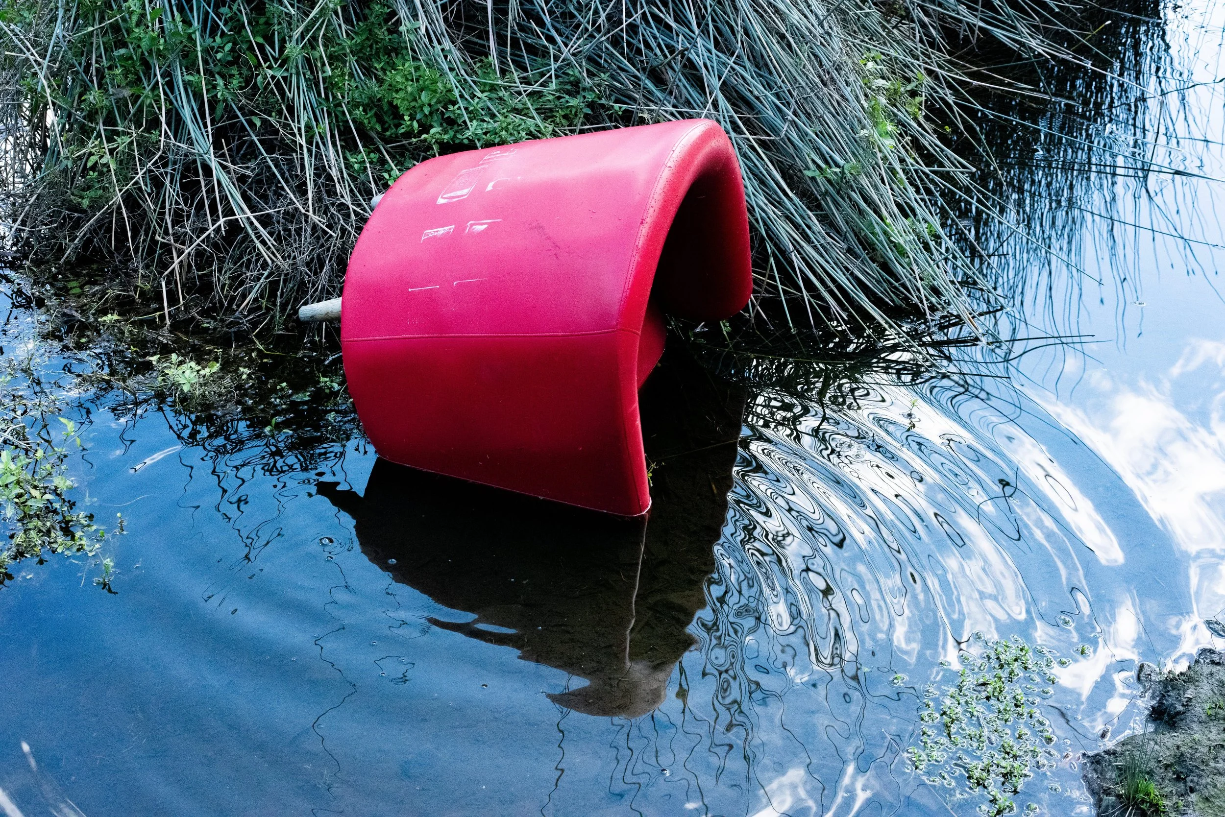 A red mailbox tilted in a body of water near the shoreline, surrounded by reeds and plants, with a lake and sky reflected in the water.