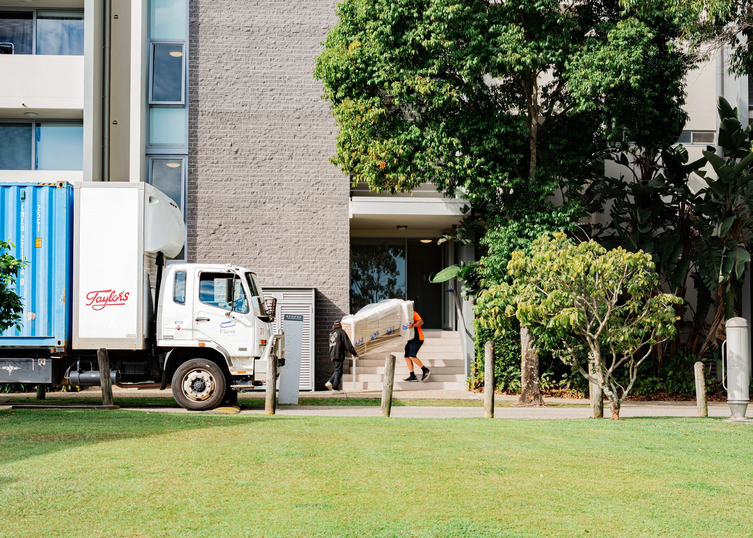 People are unloading items from a white delivery truck parked in front of a modern apartment building with a brick facade and large windows. There are trees and a grassy area in the foreground.
