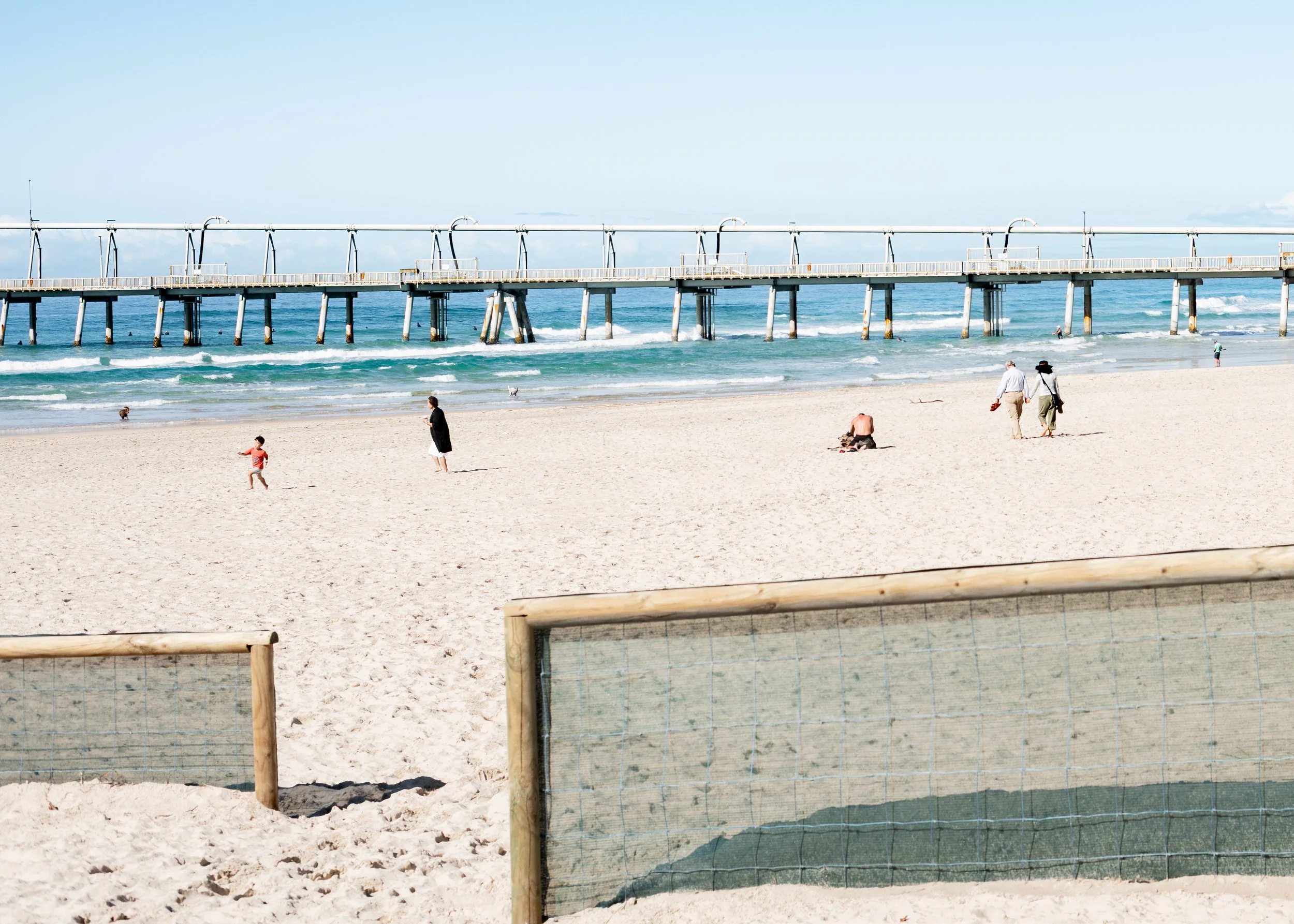 Beach with people walking and children playing, pier extending into the ocean, sandy shore, and a fence in the foreground.
