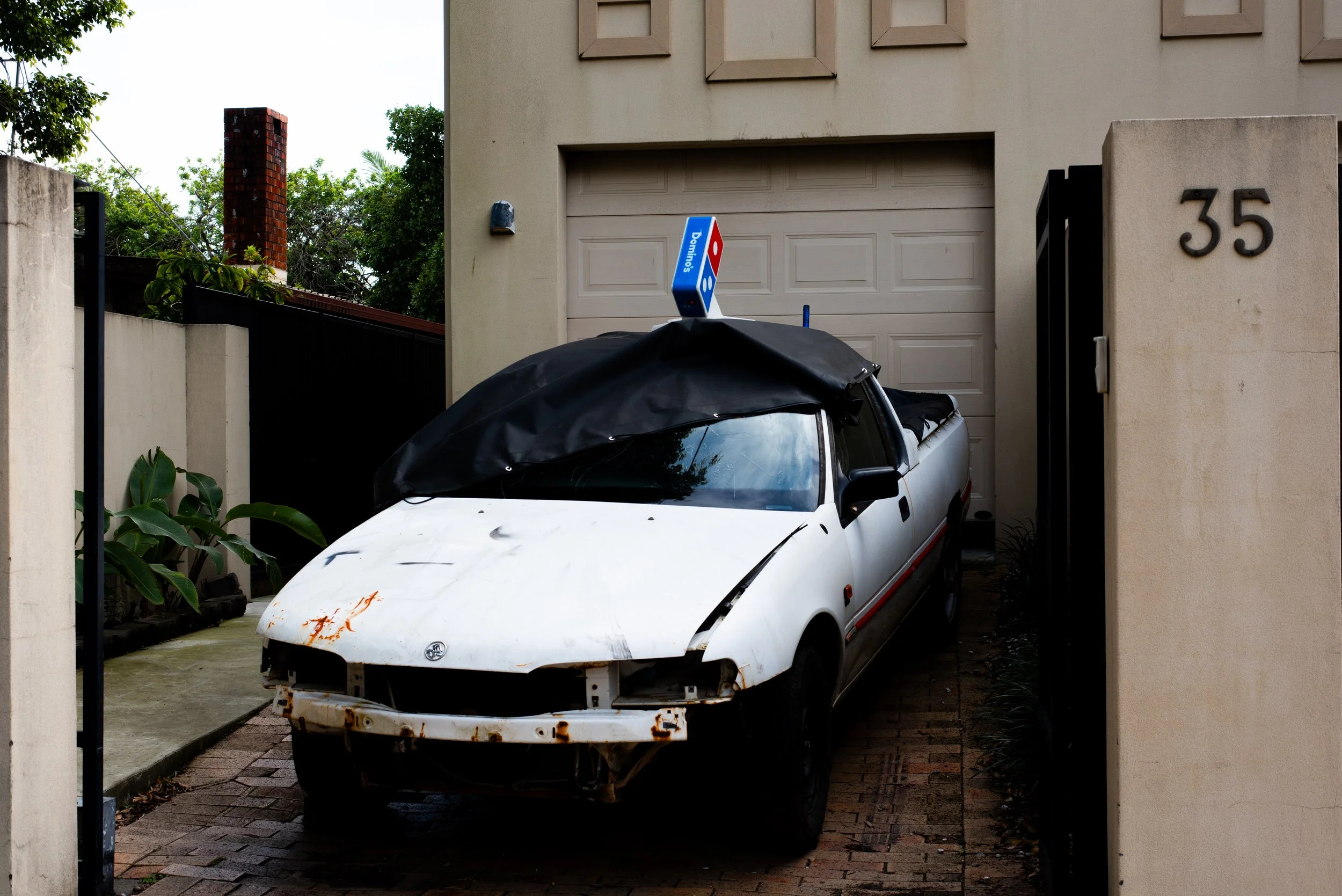 A white car with rust spots and a black cover over the back, parked in front of a garage with a Domino's sign on top, located between two stone walls with house number 35.