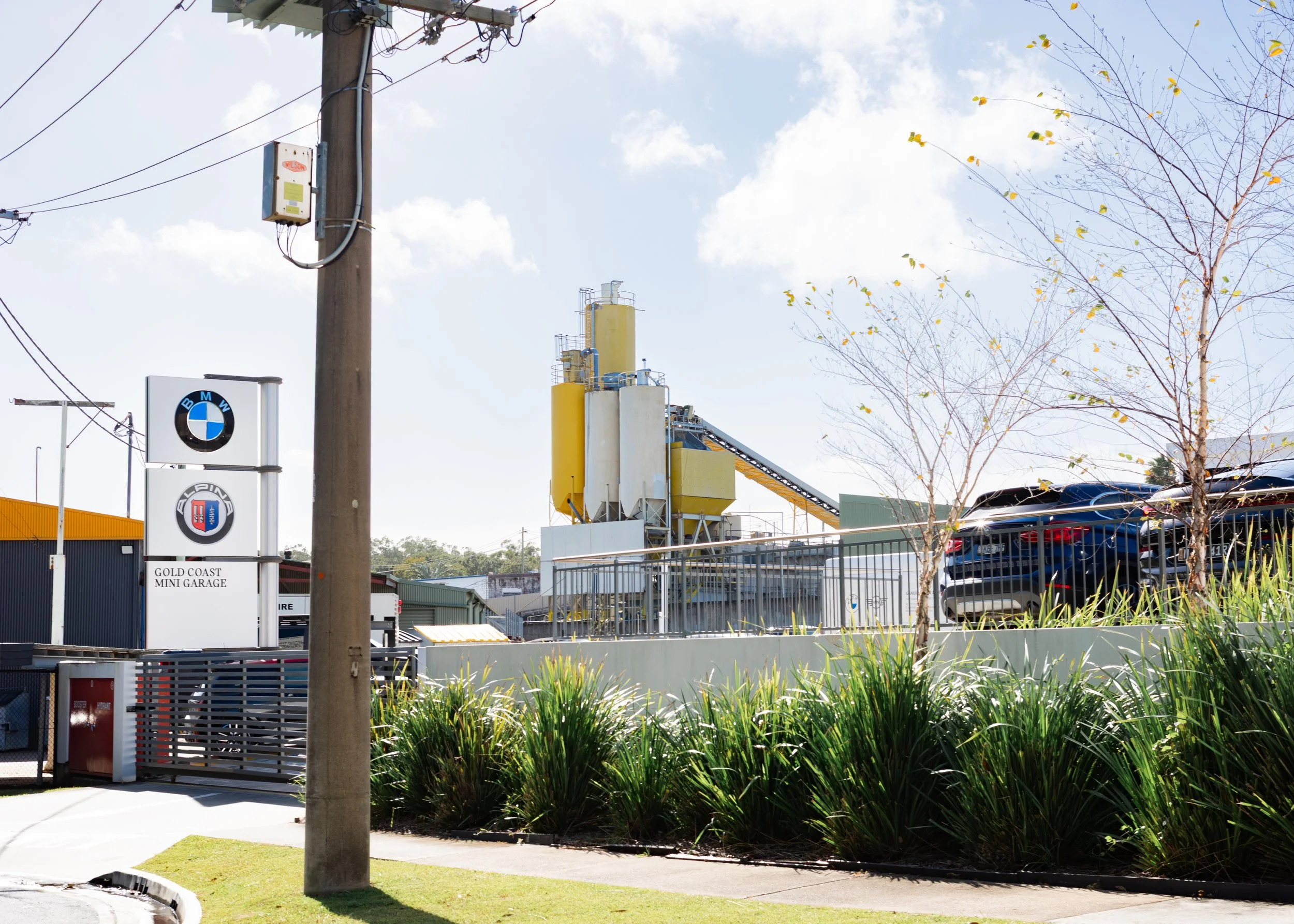 A street scene with power lines, a utility pole, and a sign for Gold Coast Mini Garage. In the background, there are industrial structures and parked cars, with a partly cloudy sky overhead.