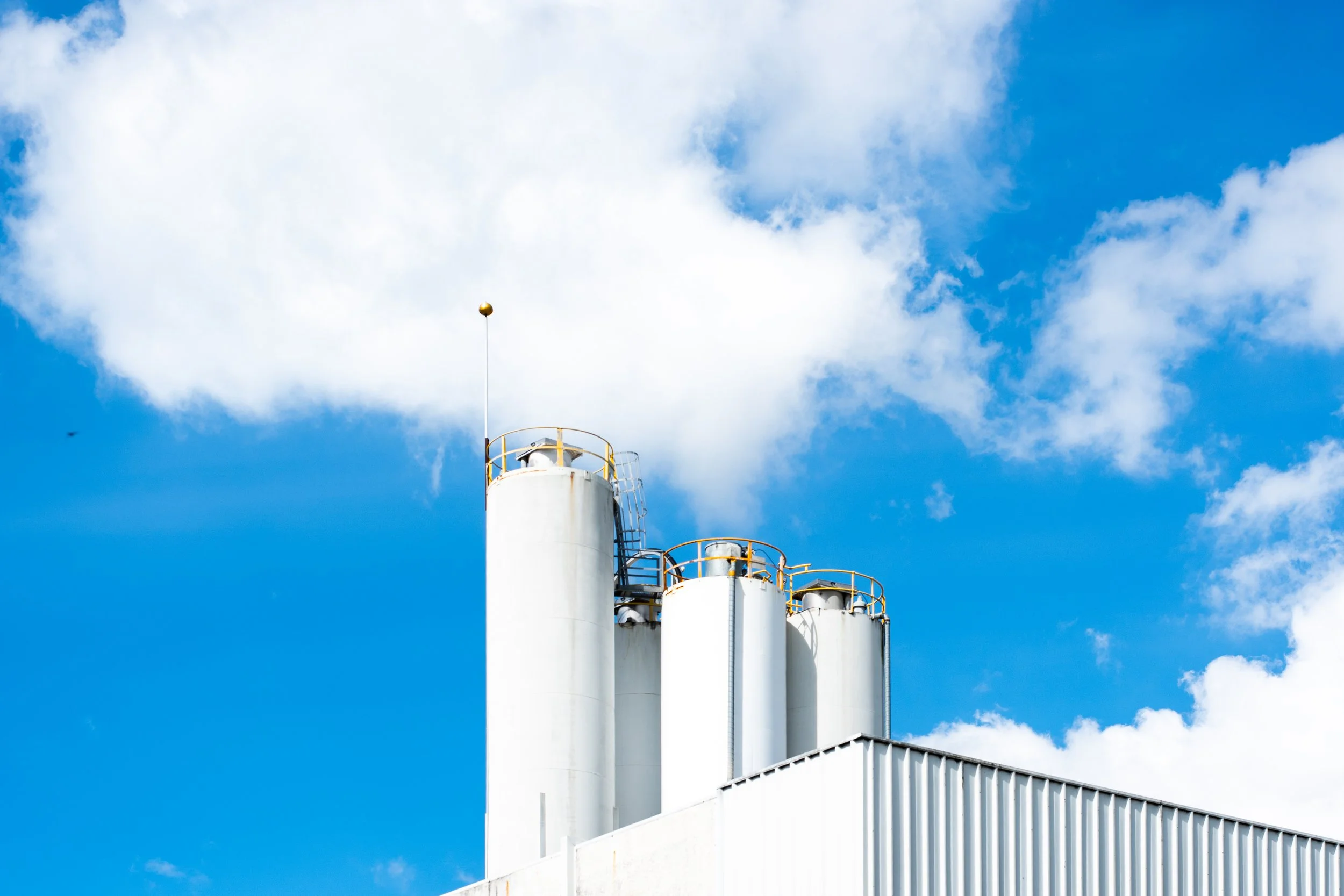 Industrial building with tall white structures and yellow safety railings against a bright blue sky with scattered clouds.