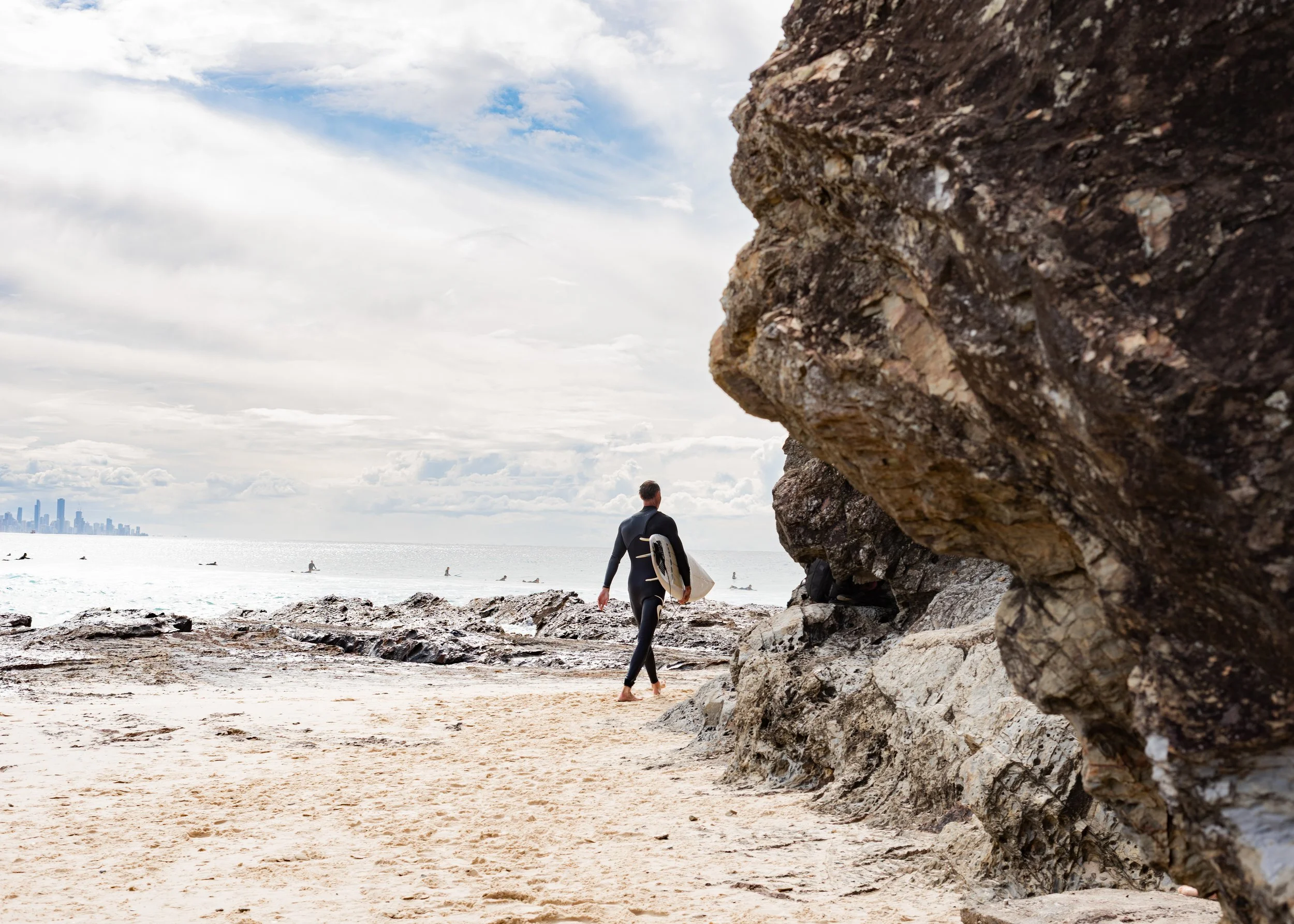 A person carrying a surfboard walking along a beach near large rocks, with the city skyline in the distance and a cloudy sky overhead.