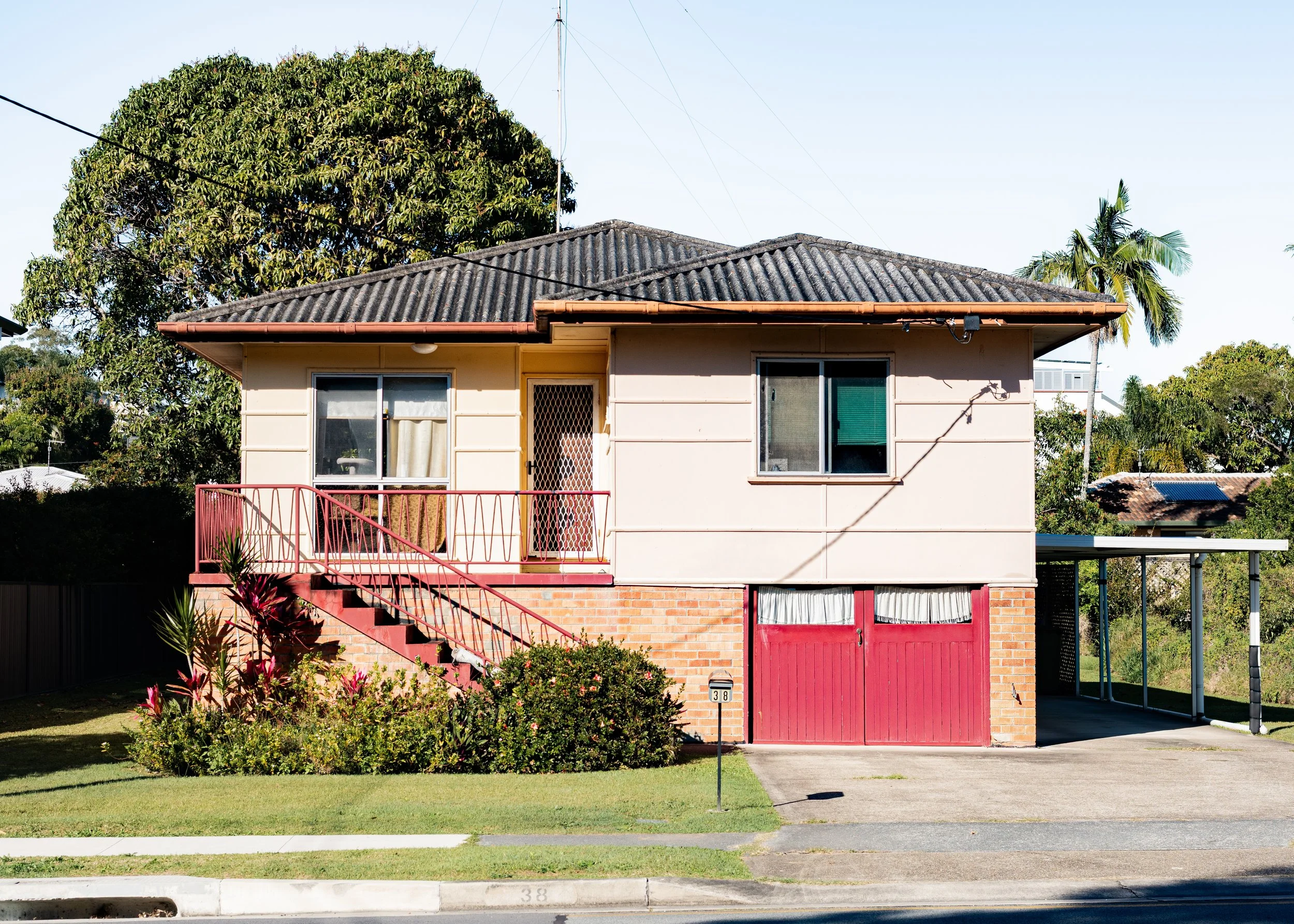 A two-story house with a beige exterior, red gate, and brick base, with a front yard landscaped with shrubs and a lawn, under a clear sky. The house has two windows and a door on the upper level, accessible by red stairs, and features a red railing. 