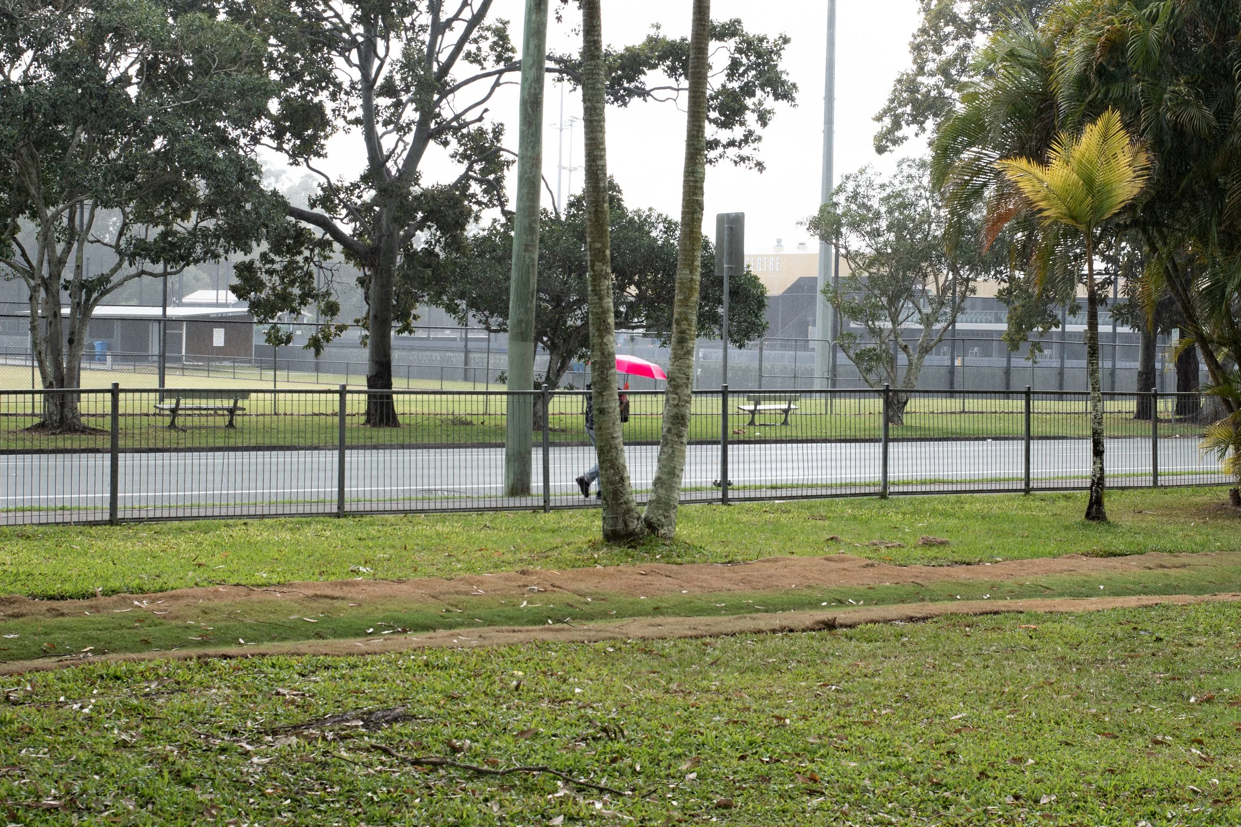 A rainy day scene at a park with green grass, trees, a black metal fence, and a person holding a red umbrella walking along the sidewalk.