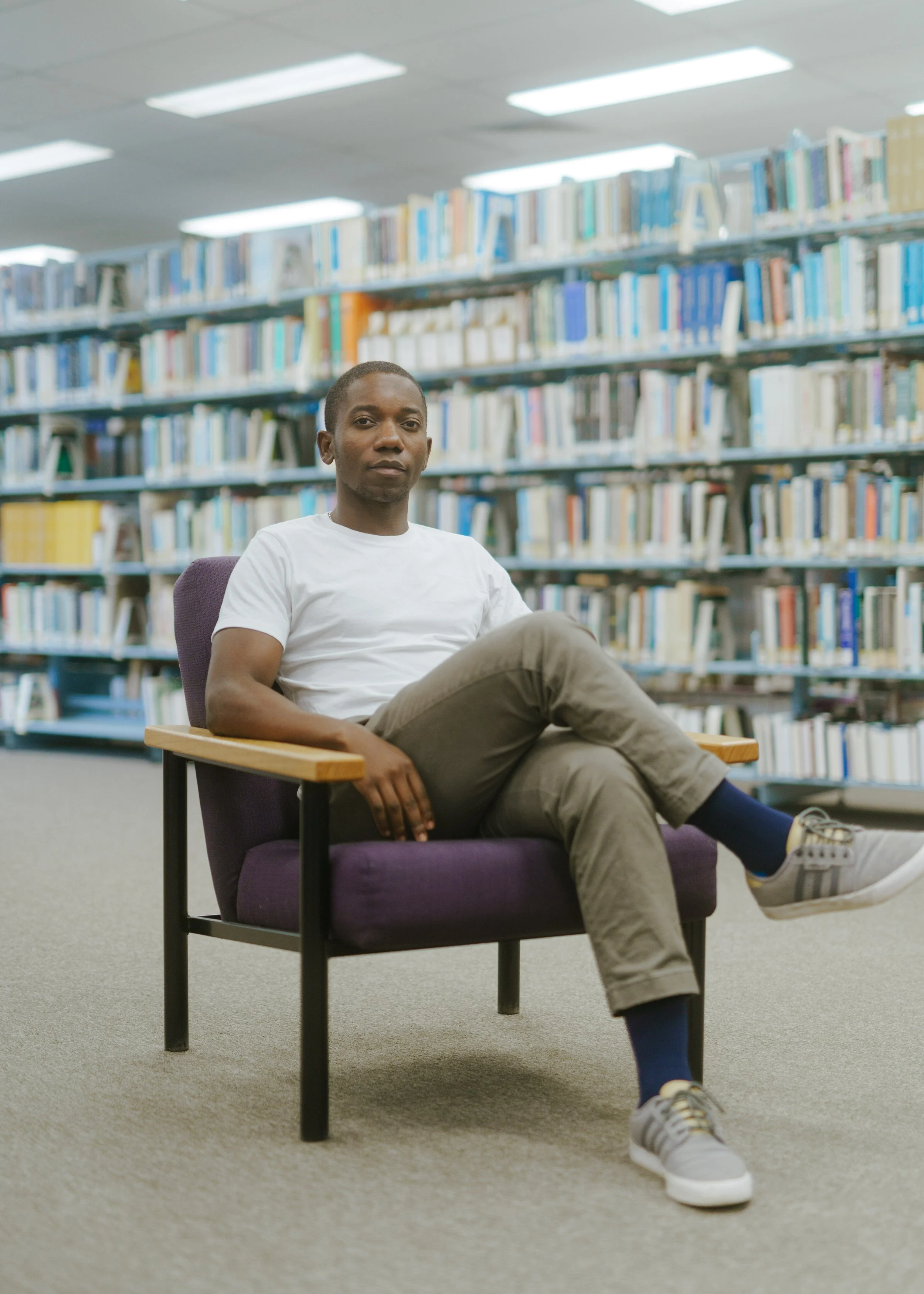 A young man in a white t-shirt and khaki pants sitting in a purple armchair in a library, with shelves of books in the background.