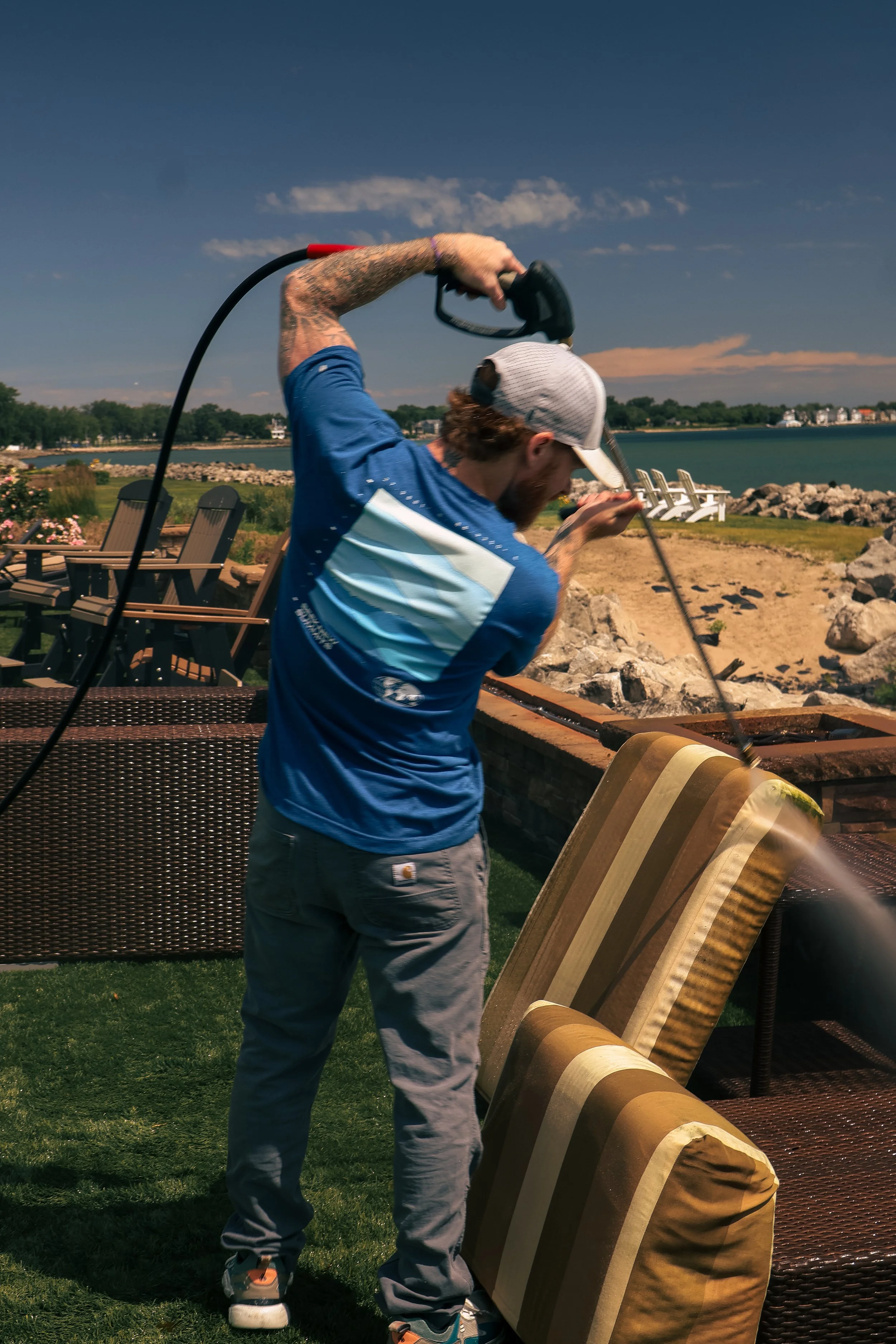 Man in blue shirt and gray cap hammering a large cardboard box outside near water, with chairs and rocks in the background.