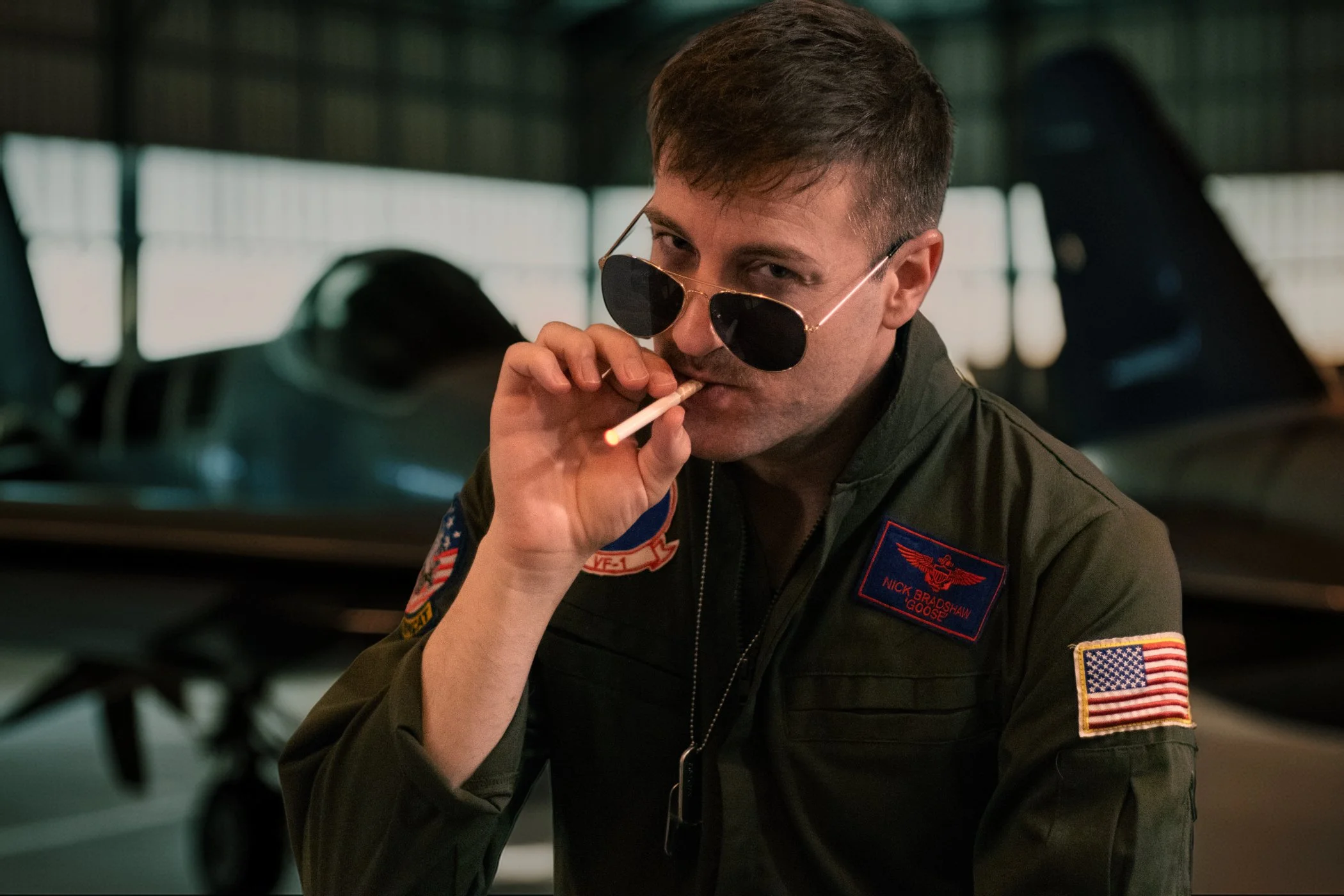 A man in a military flight suit with patches, wearing sunglasses, holding a cigarette to his lips indoors with a blurred airplane in the background.