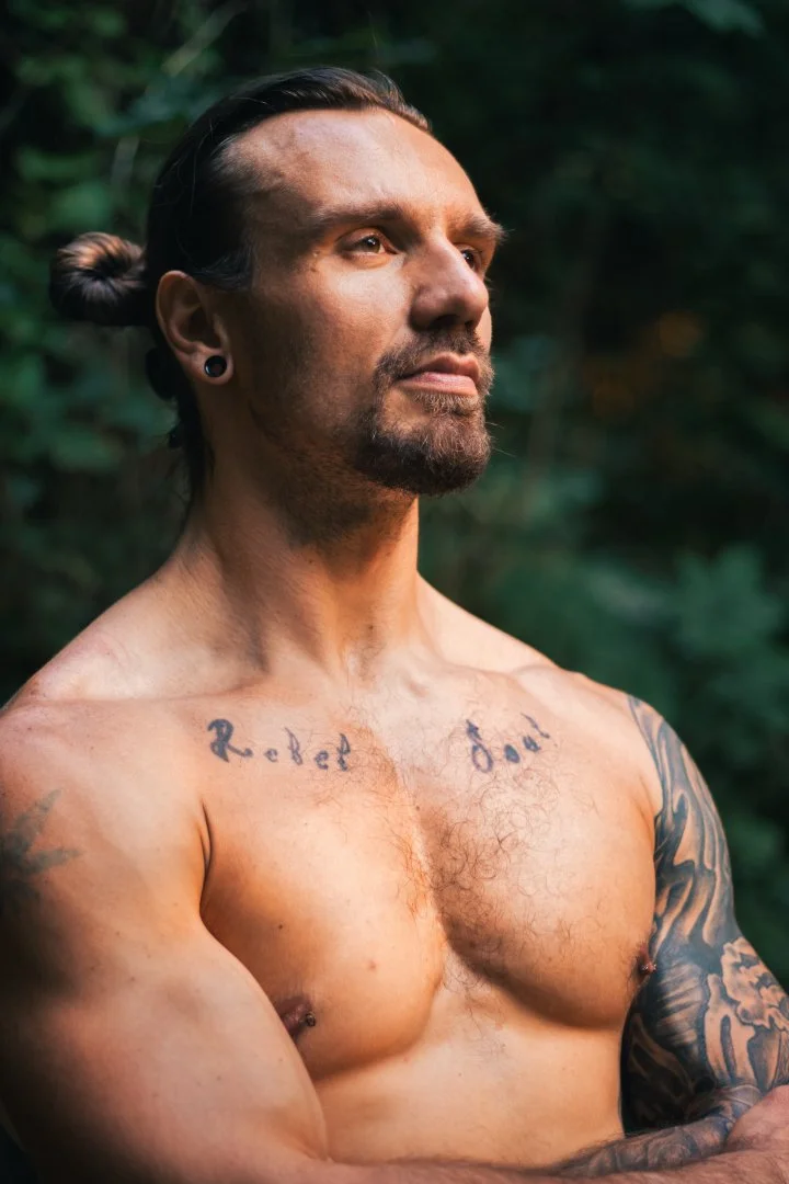 A shirtless man with tattoos on his chest and arms standing outdoors with a blurred green background, looking serious and contemplative.