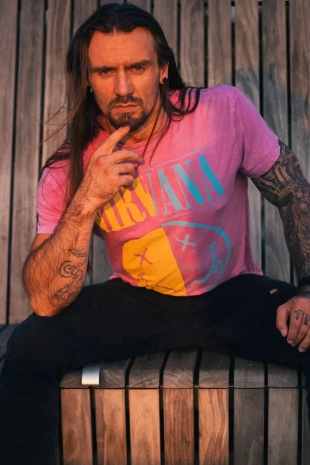 A man with long dark hair and tattoos, wearing a pink Nirvana t-shirt, sitting on a wooden bench against a wooden fence, making a thoughtful pose with his hand on his chin.