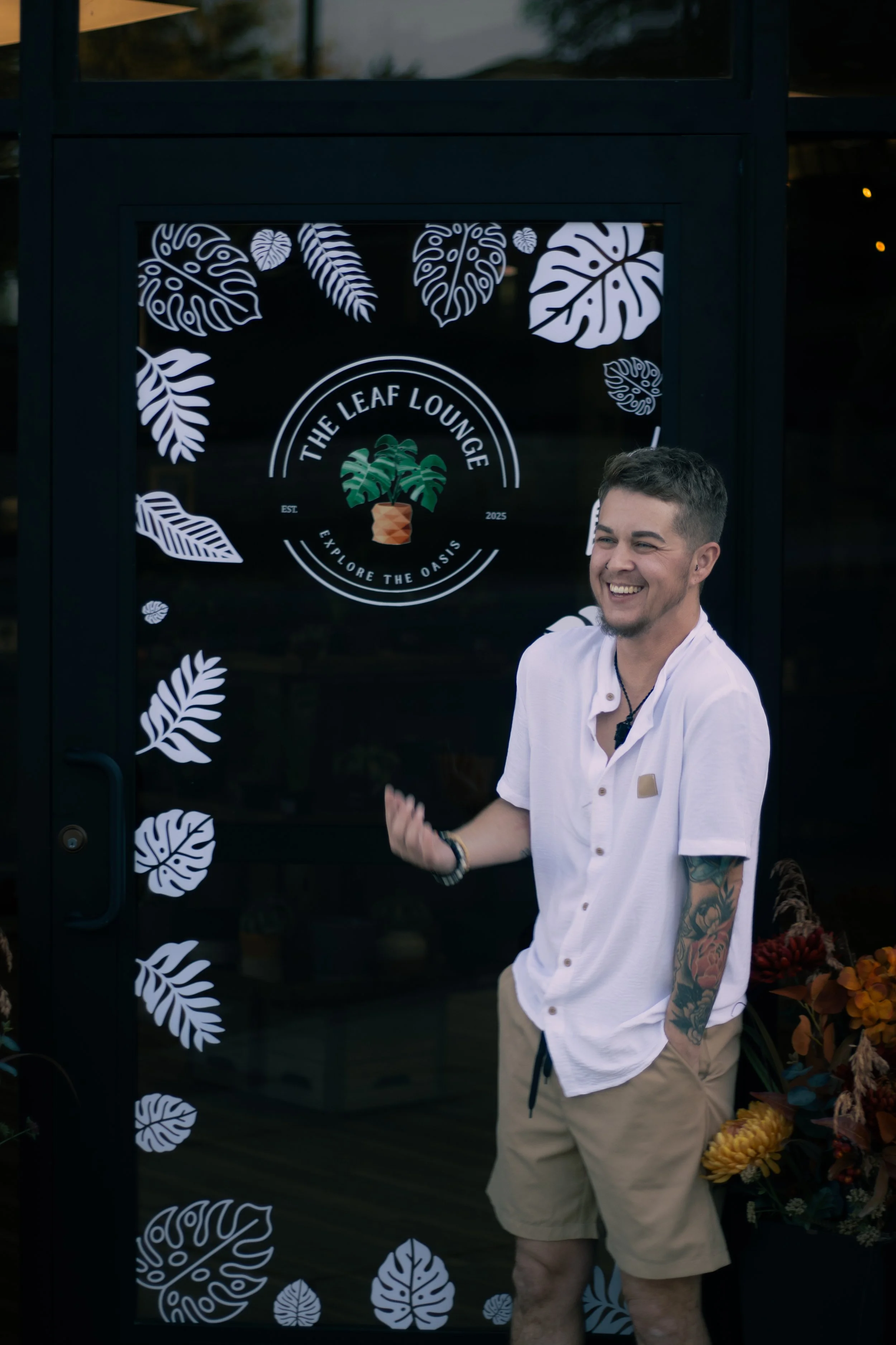 A smiling man with short dark hair and tattoos on his arm, wearing a white short-sleeve shirt and beige shorts, standing outside in front of a black door with white monstera leaf designs. The door has a sign for 'The Leaf Lounge' with the tagline 'Ex