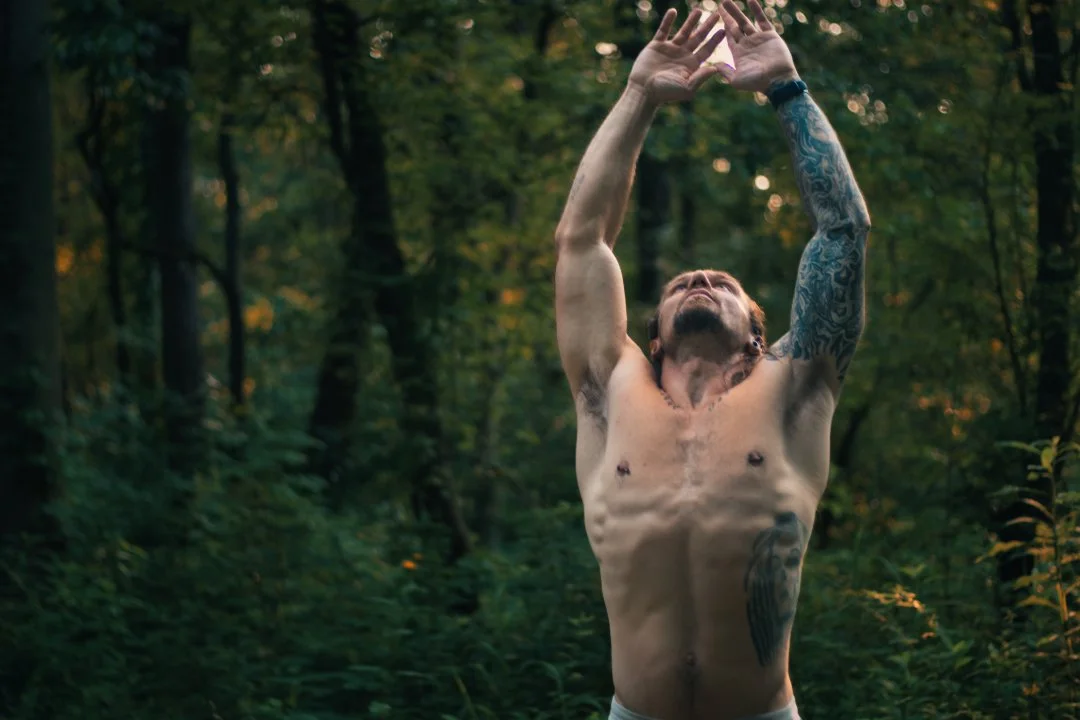 A shirtless man with tattoos on his arms and torso standing in a forest, reaching upward with his hands raised, gazing toward the sky.