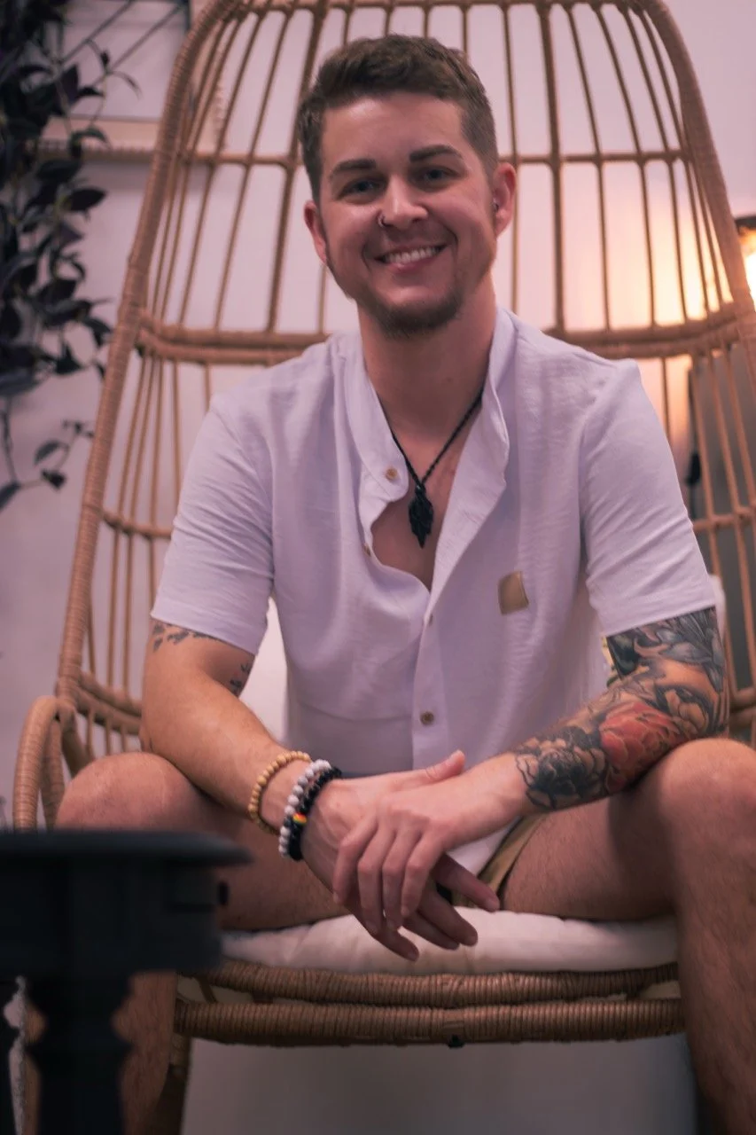 A young man with short brown hair and tattoos on his arms, smiling, sitting cross-legged in a wicker chair indoors with warm lighting and plants nearby.
