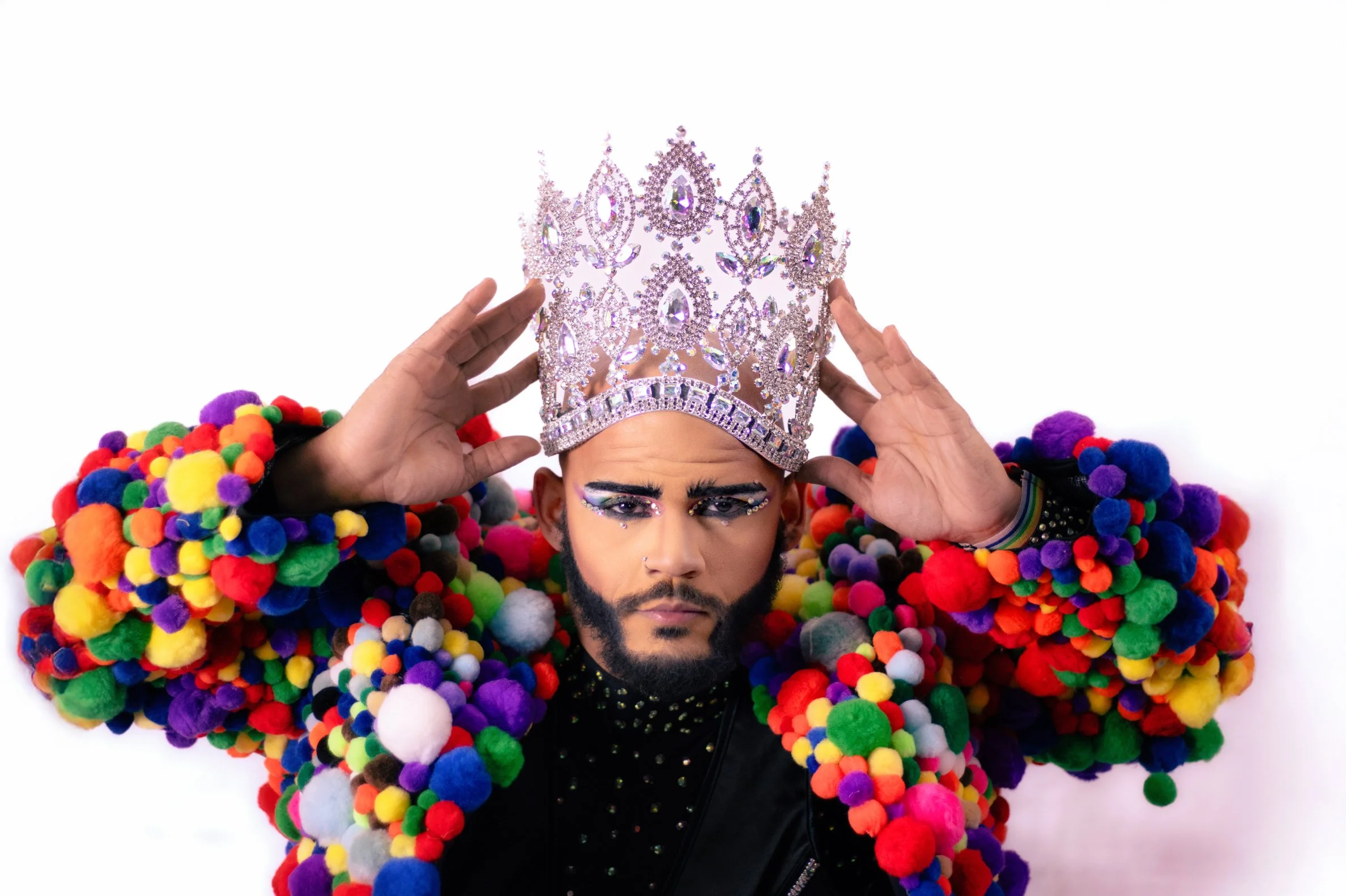 Person with colorful pom-pom cape and a large, ornate crown holding it with both hands.