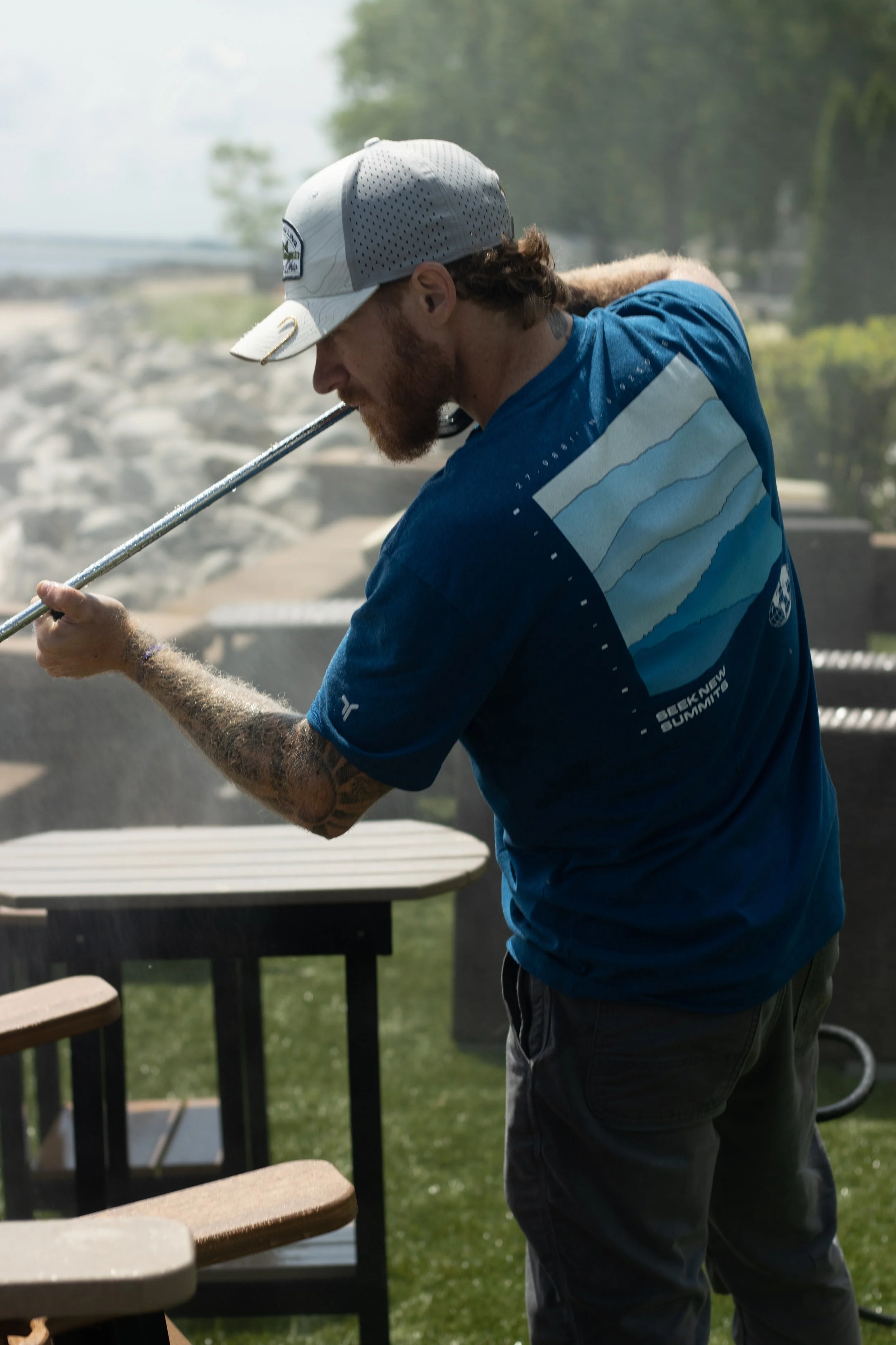 A man with a beard and tattoos wearing a gray cap and blue shirt playing mini golf, preparing to hit a golf ball with a club outdoors on a sunny day.