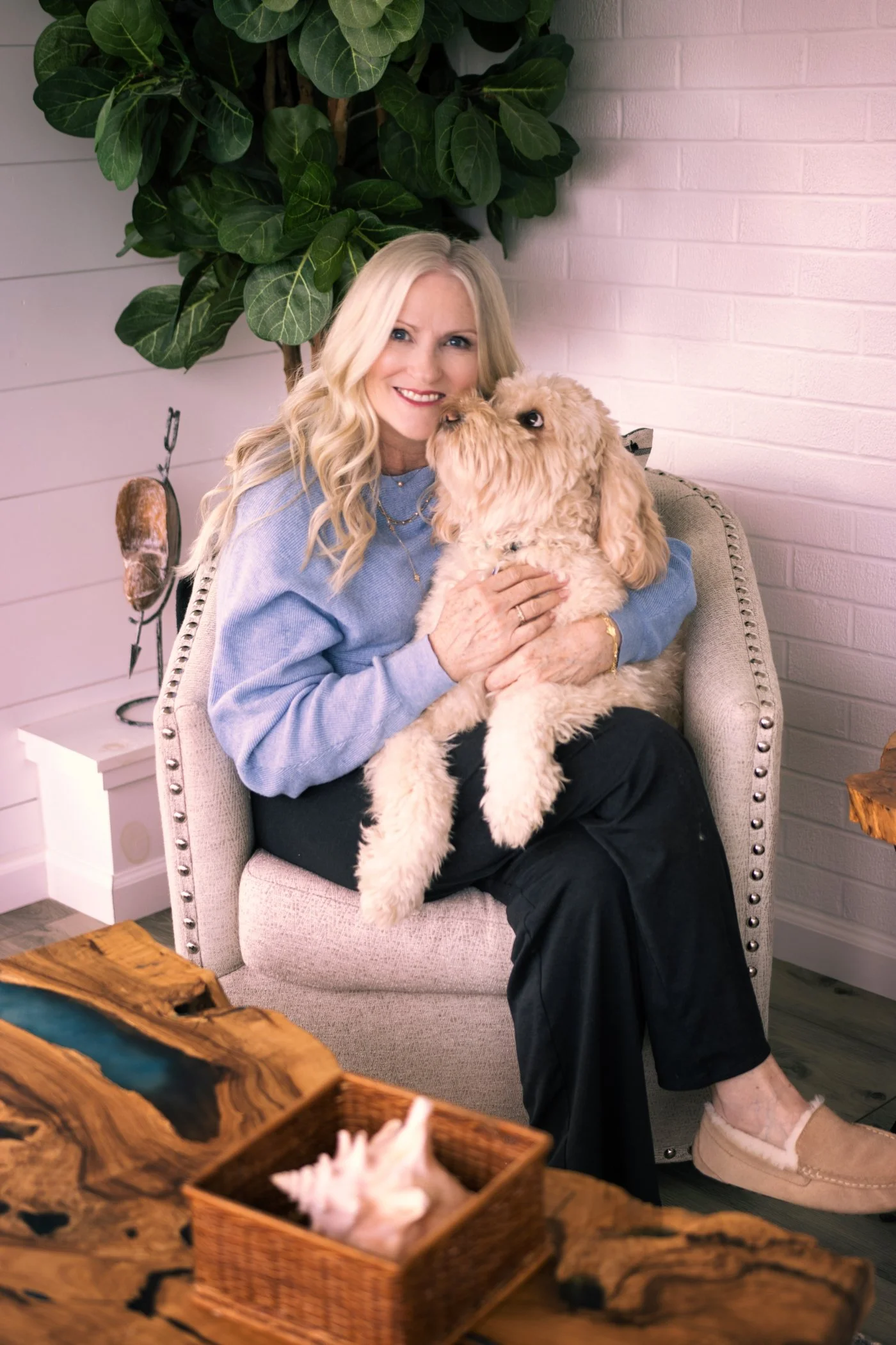 A woman with blonde hair smiling while holding a fluffy tan dog in a cozy indoor setting. The woman is wearing a light blue sweater and black pants. The background includes a white brick wall and a large green potted plant.