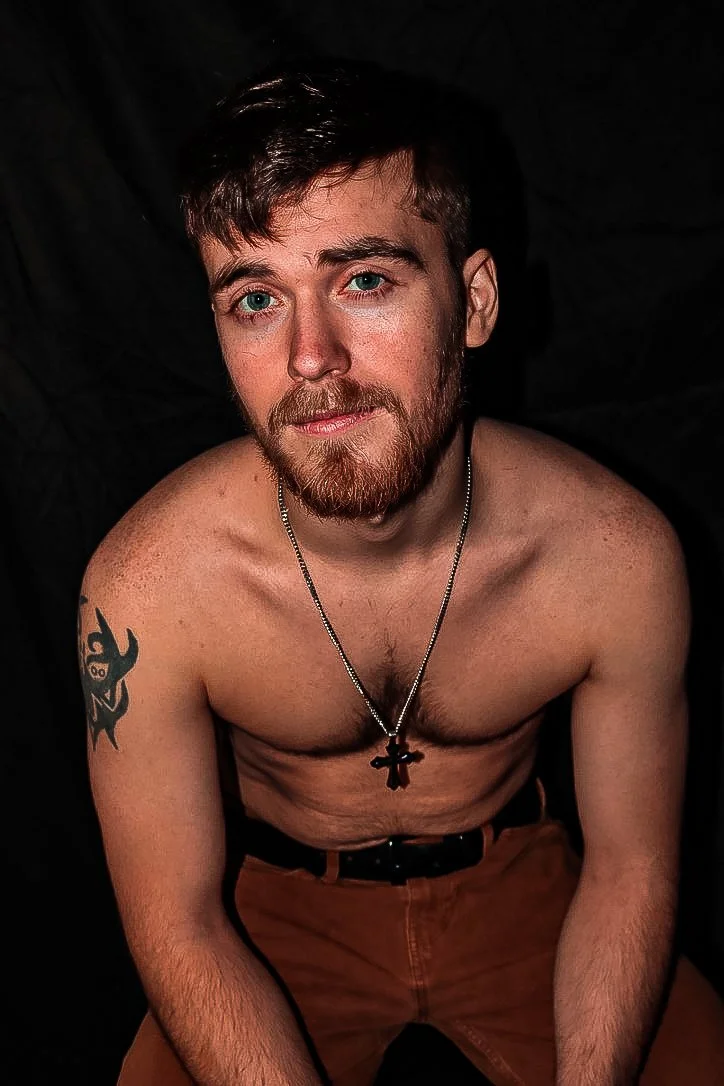 A shirtless young man with red hair and a beard, wearing a cross necklace, sitting against a black background.